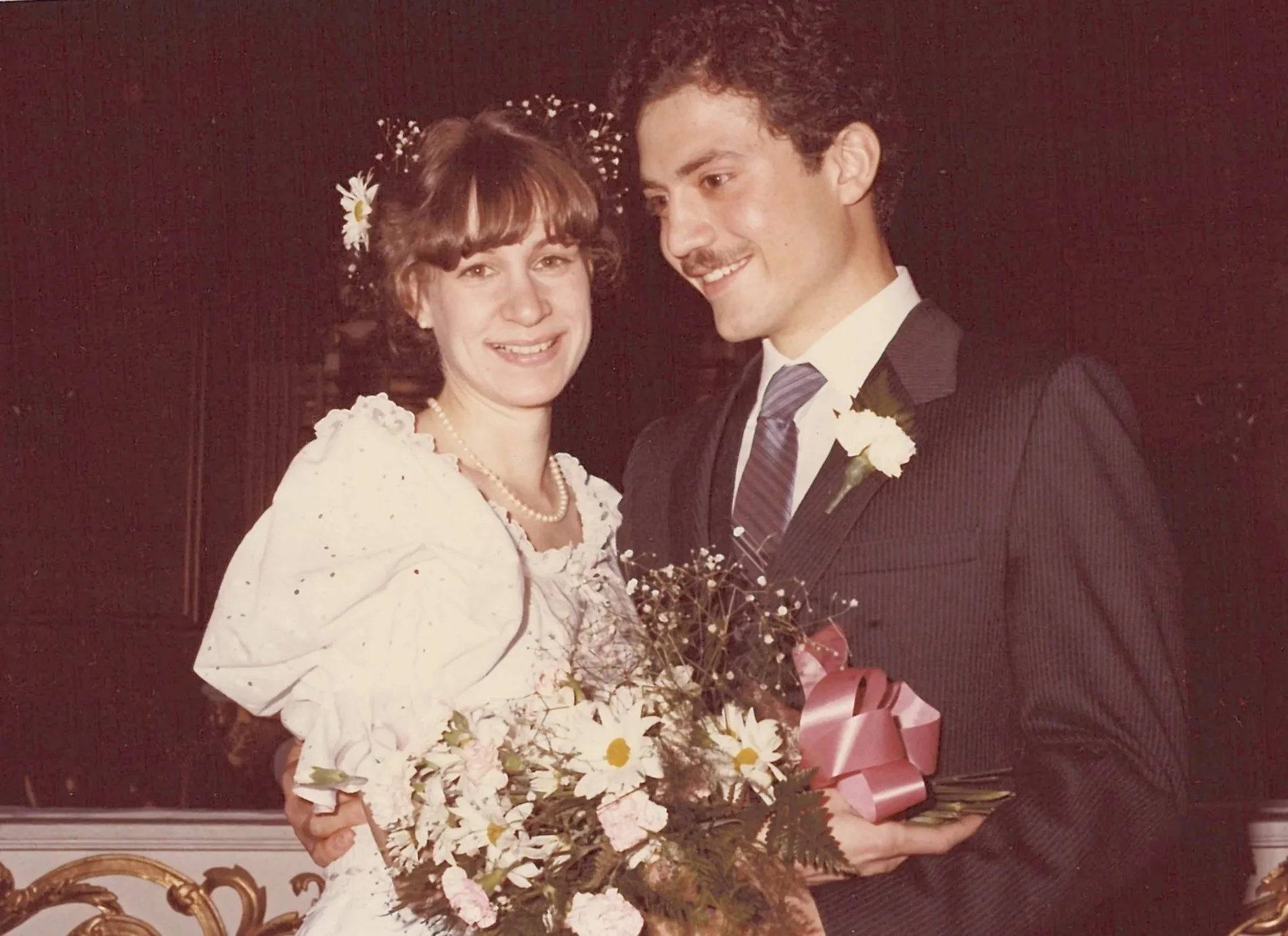 A young woman in a white dress and pearl necklace holding a bouquet of flowers, standing next to a young man in a dark suit and tie, both smiling, at what appears to be a wedding or special event.