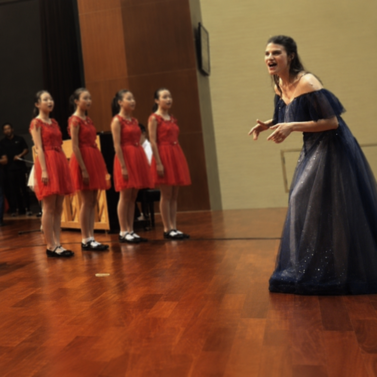 A woman in a dark blue, sparkly gown appears to be conducting or speaking to four young girls in matching red dresses with lace tops, who are standing in a line on a wooden stage or floor.