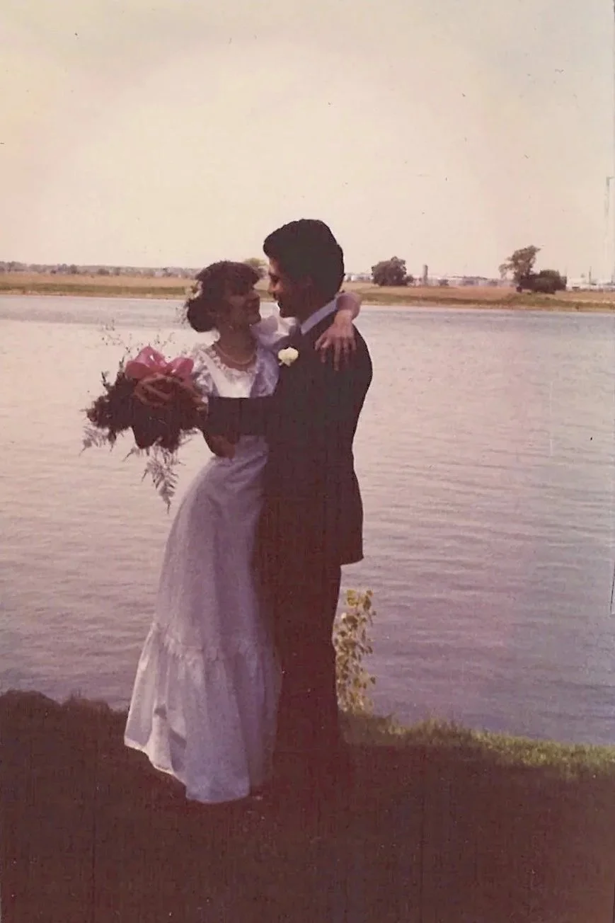 A couple in wedding attire embracing by a body of water, with the woman holding a bouquet of flowers and the man in a suit wearing a boutonnière.
