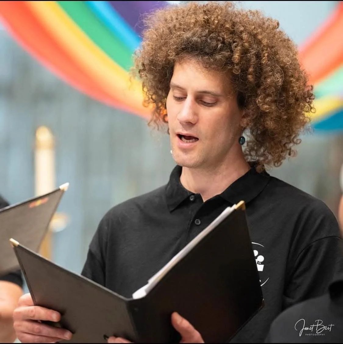 A person with curly hair wearing a black shirt, holding a black folder and singing or reading from it, with a colorful rainbow banner in the background.