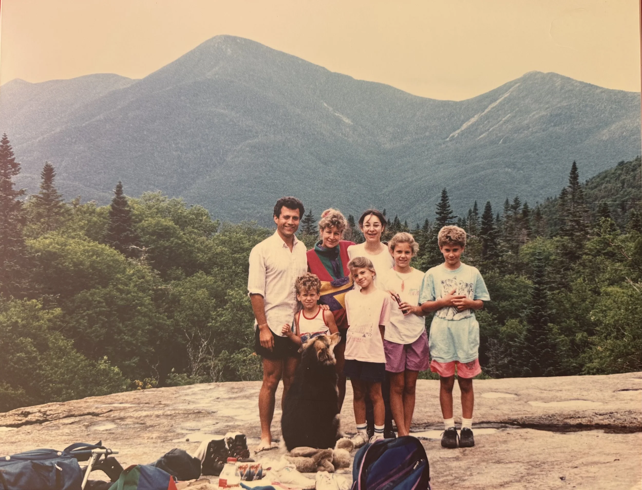 Group of eight people, including children and adults, standing on a rocky outdoor area with a forest and tall mountains in the background, likely on a hiking trip.