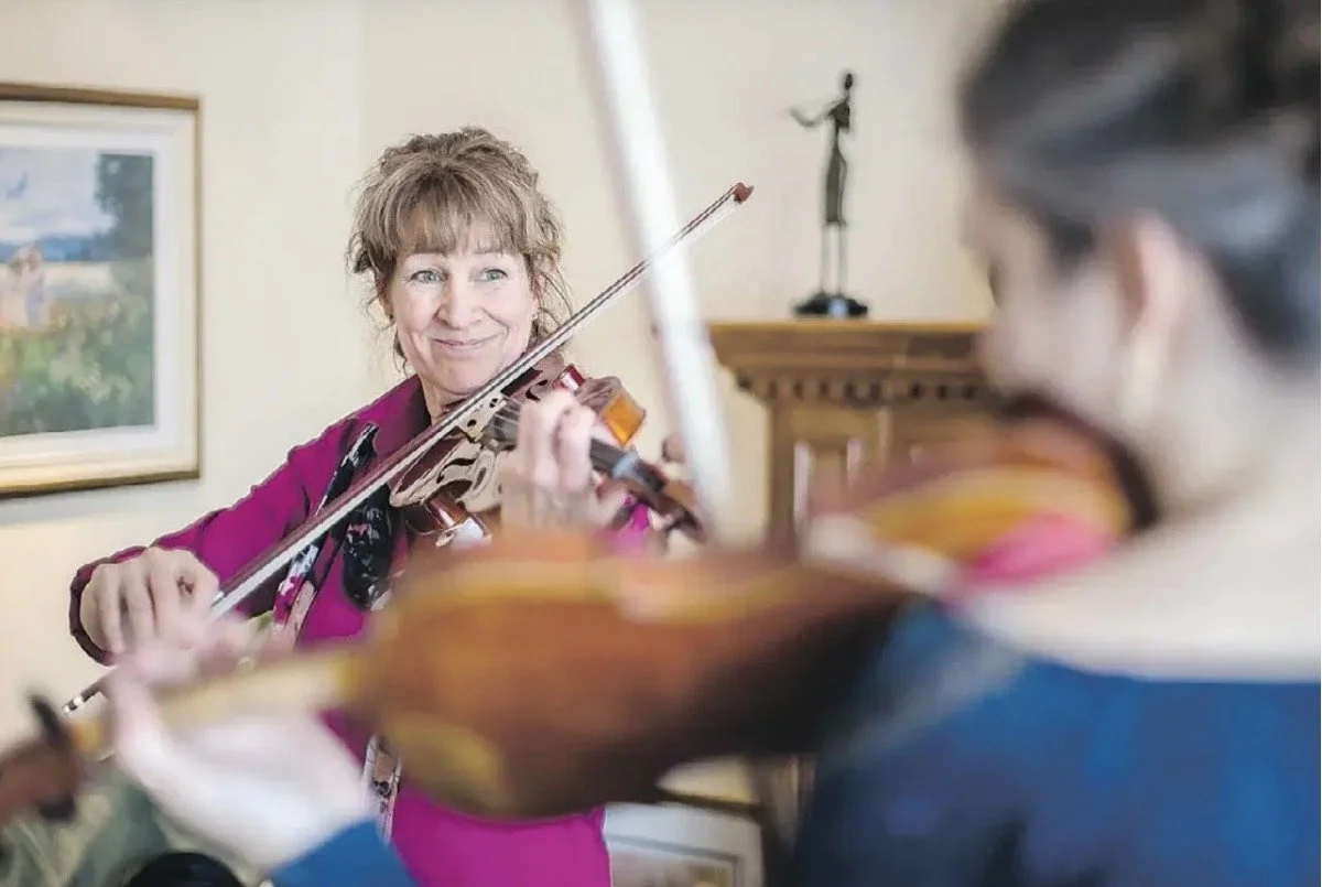 A woman with shoulder-length hair, wearing a pink jacket, playing a violin and smiling at a young girl also playing a violin.