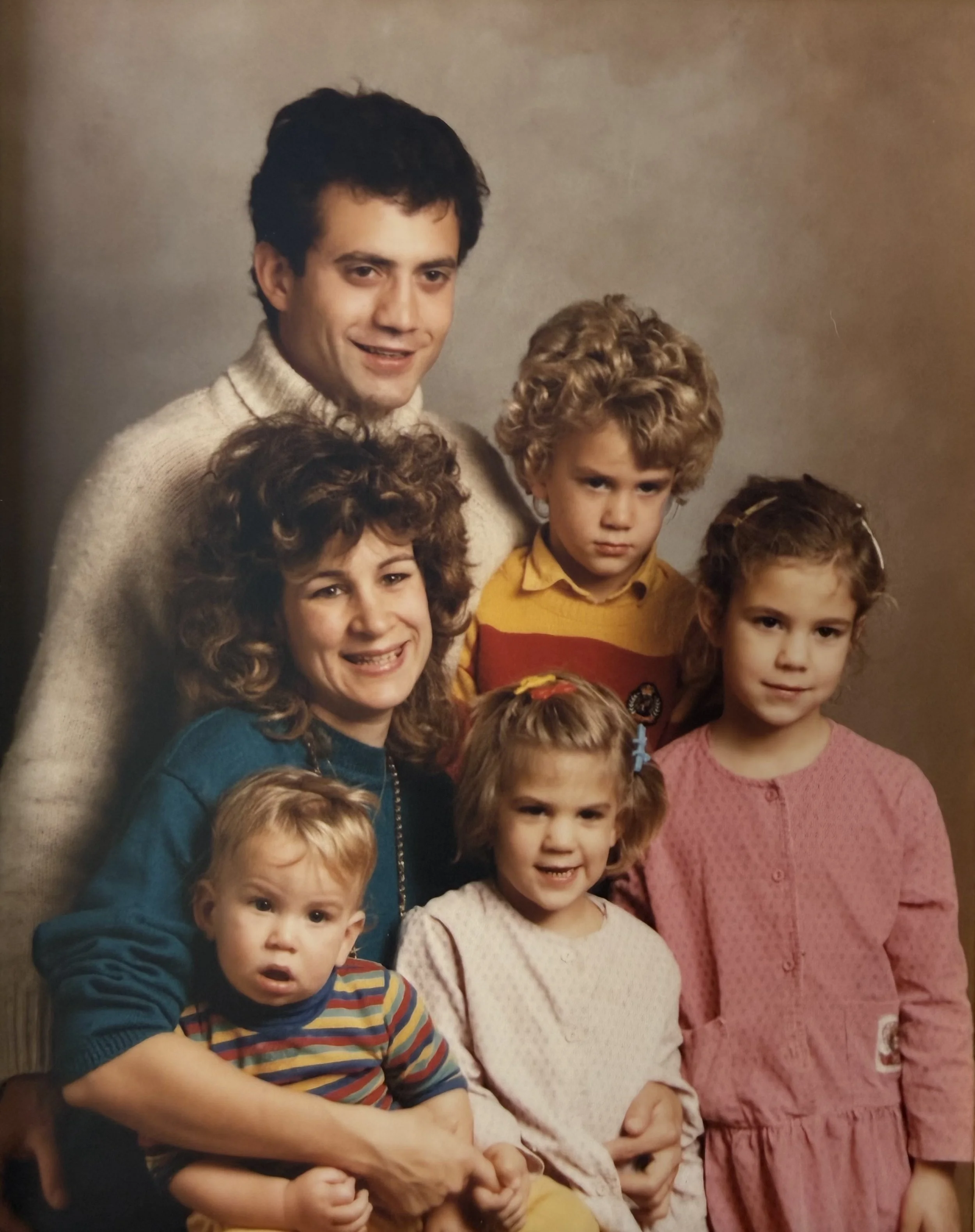 A group photo featuring a man with dark hair and light skin wearing a cream turtleneck, four girls with curly hair and light skin, dressed in colorful sweaters and dresses, standing against a beige background.