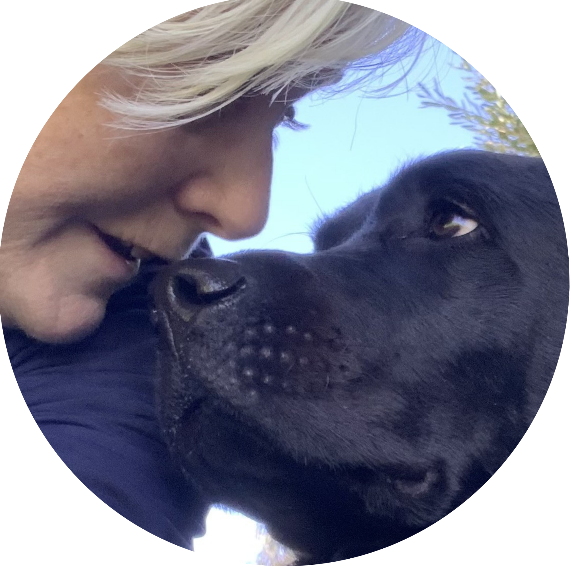 A person with blonde hair and tan skin is close to a black dog with curly fur, outdoors with trees and a blue sky