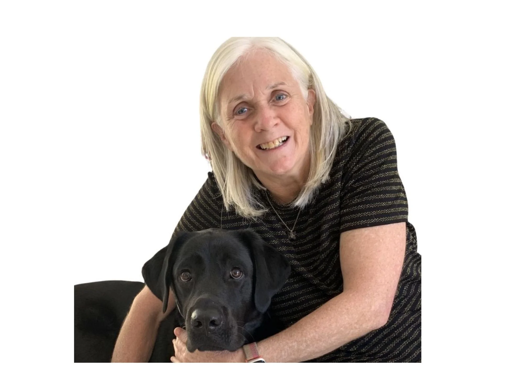 A lady with white hair smiling while hugging a black Labrador Retriever puppy.