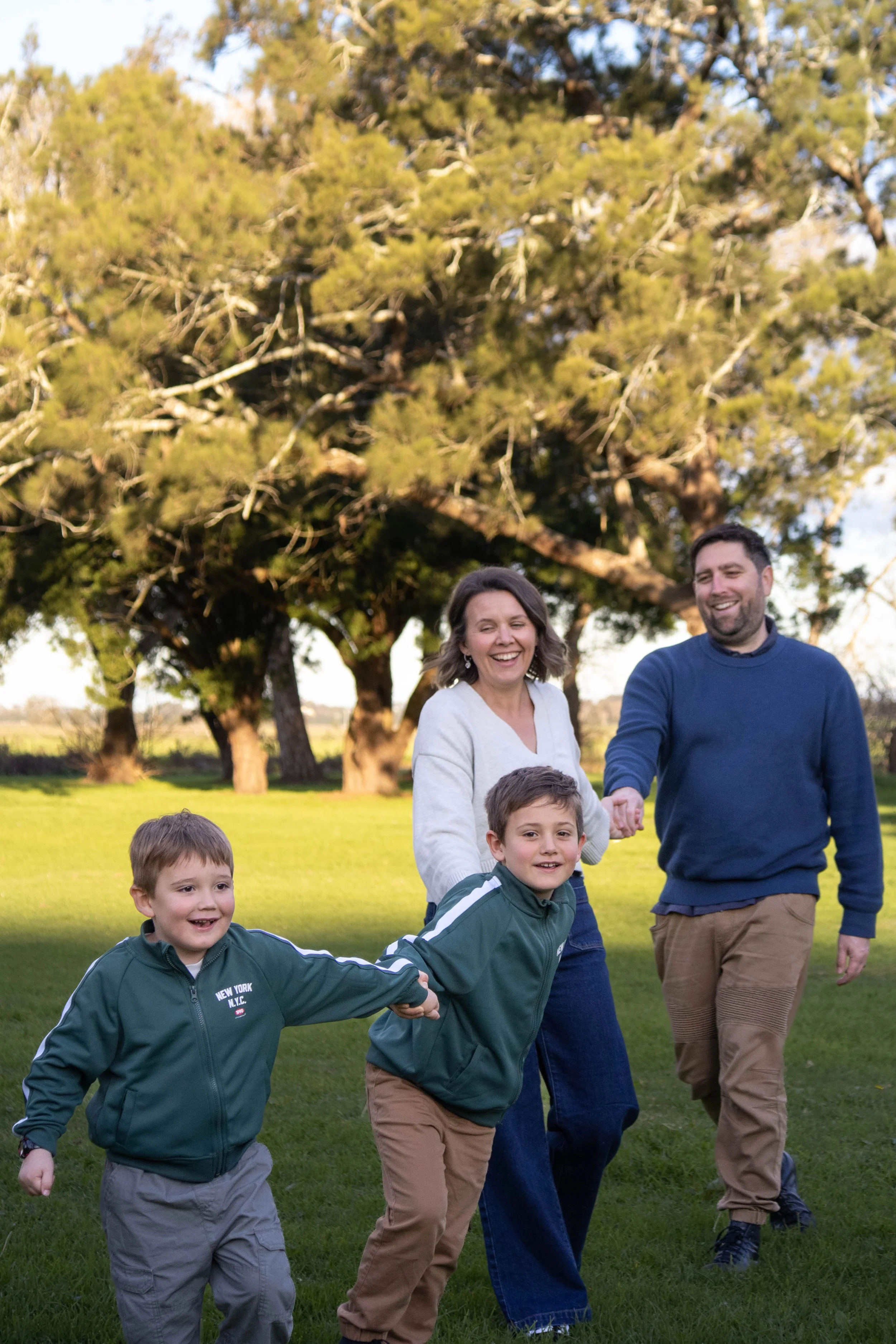 A family of four, two children and two adults holding hands and running together outdoors on a grassy field, with large trees in the background, during daytime.