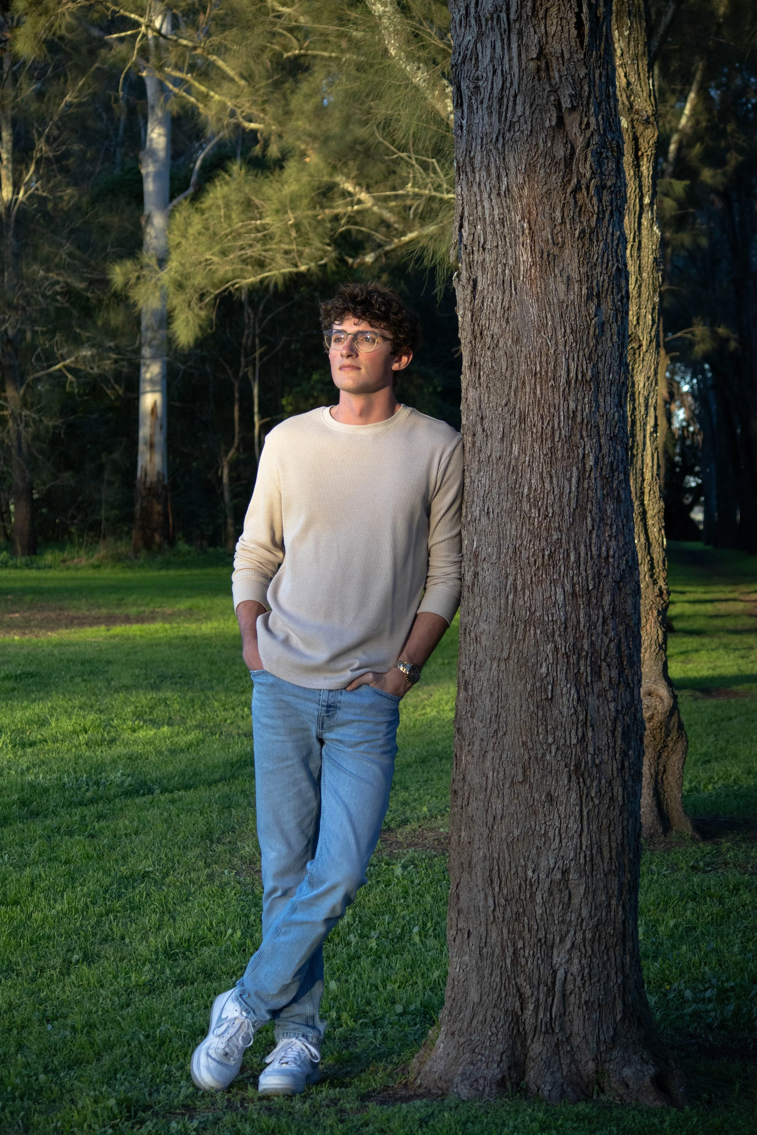 A young man with curly hair and glasses leaning against a tree in a park during daytime, wearing a beige sweater and light blue jeans, with grass and trees in the background.