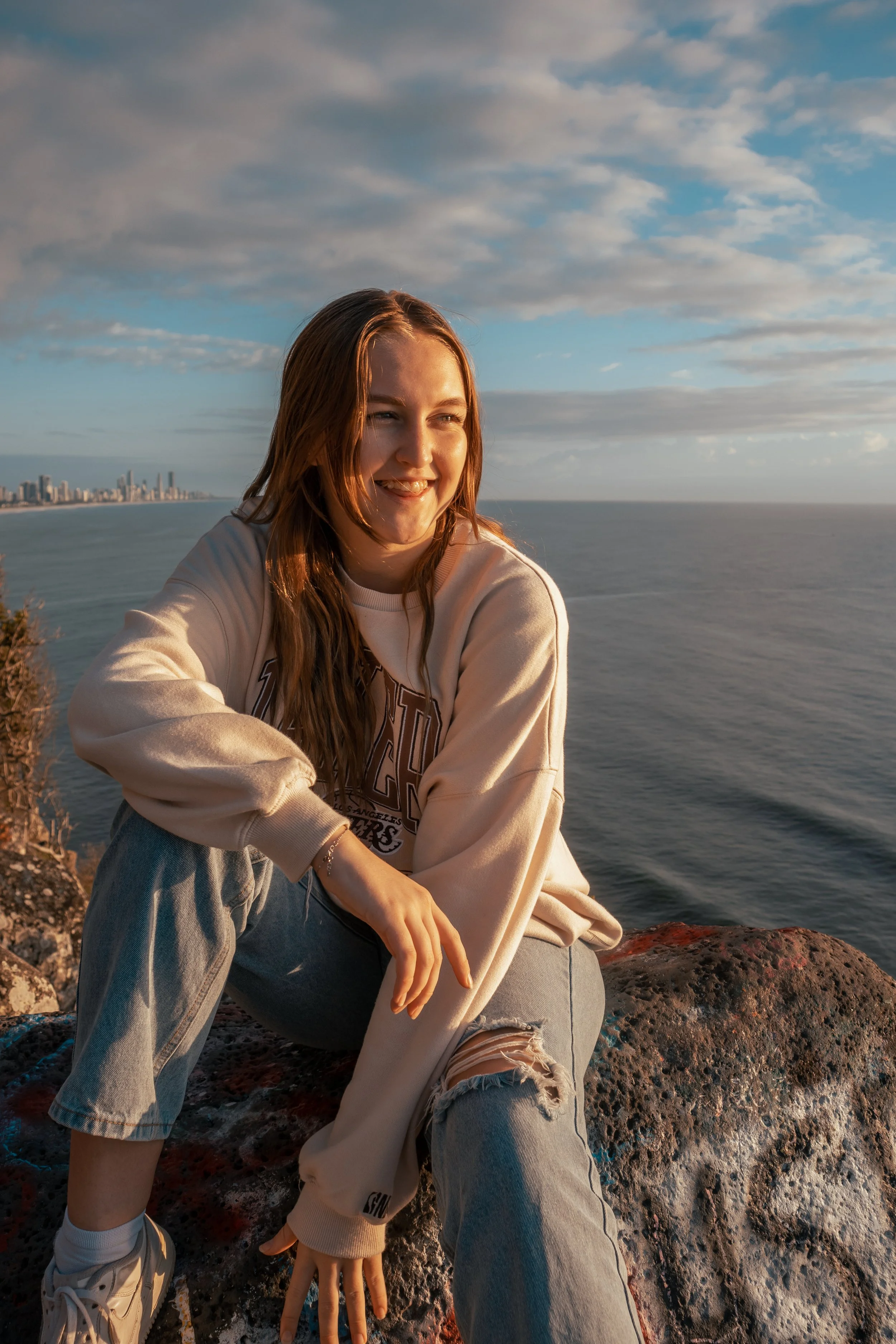 A young woman with long blonde hair smiling while sitting on a rocky surface near the ocean with a city skyline in the distance and a partly cloudy sky.