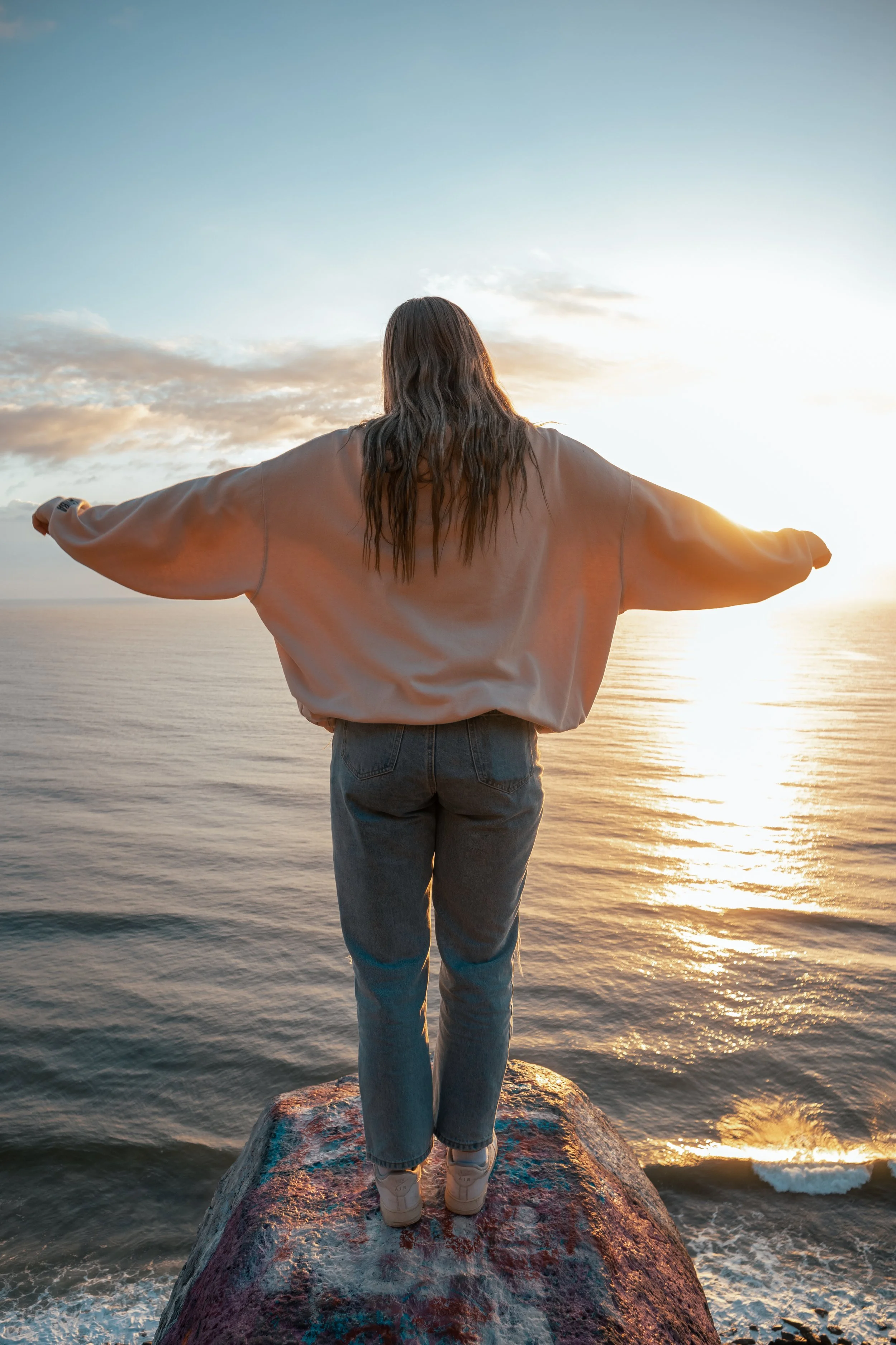 Person standing on a large rock at the edge of the water, facing the sunset over the ocean, arms outstretched, with a partly cloudy sky.