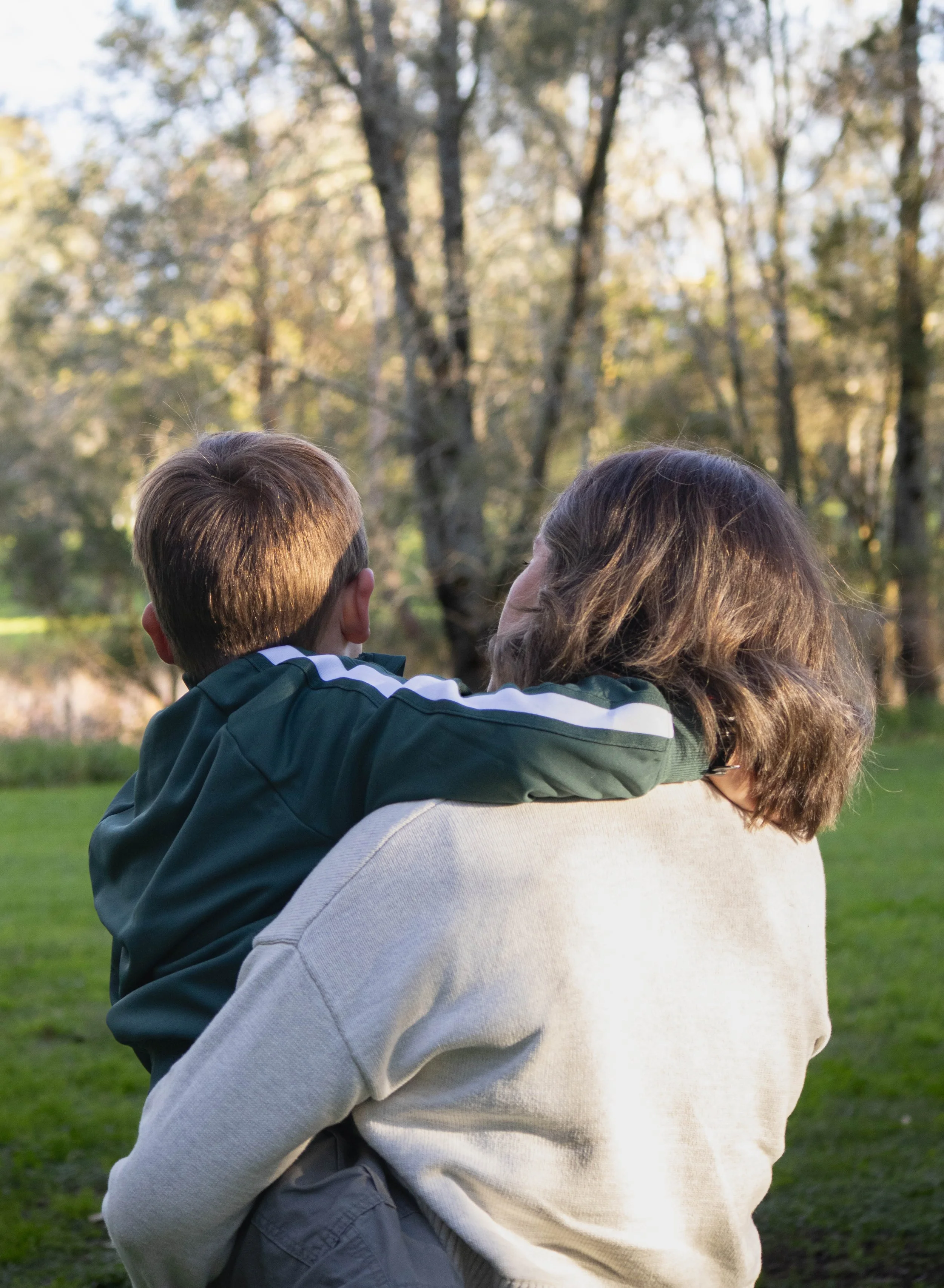 A mum carrying her son outdoors in a park, watching the trees, during sunset.