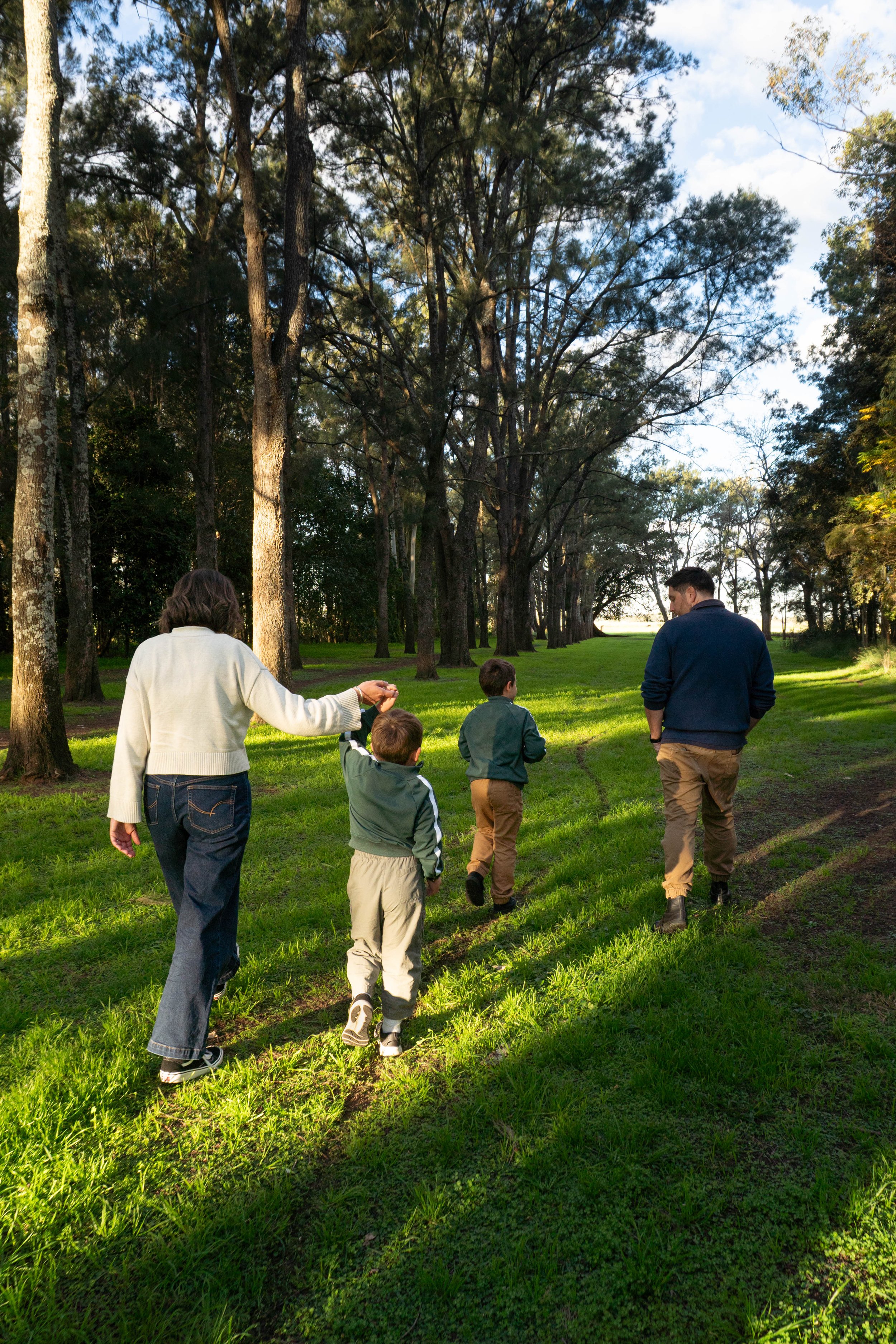 A family, including children and adults, walking in a lush green park with tall trees on a sunny day.