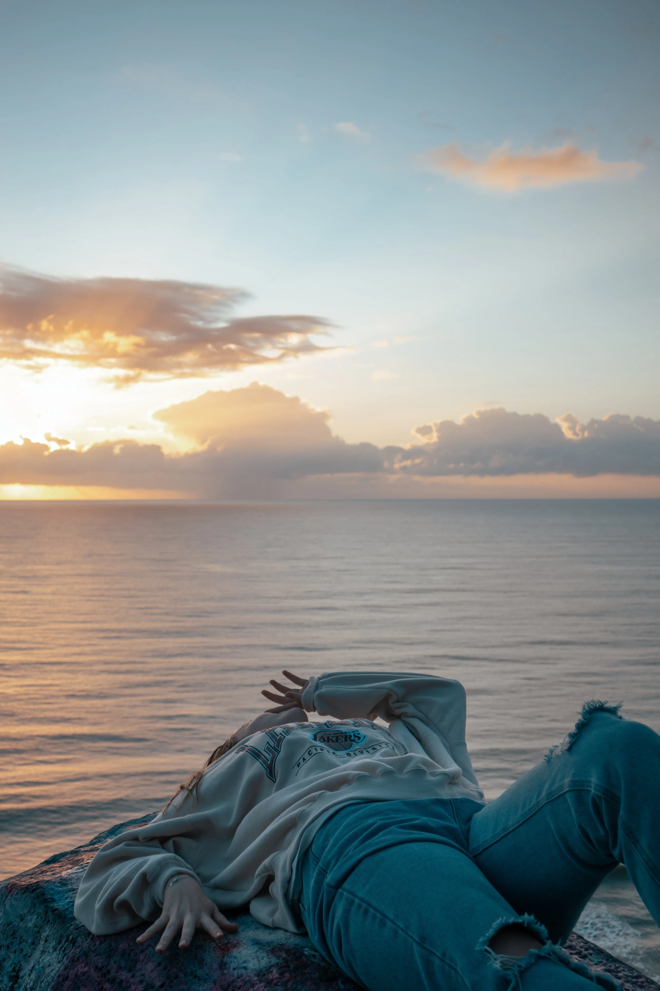 A young woman lying on a large rock by the ocean during sunrise, wearing a beige sweatshirt and ripped blue jeans, with clouds and the rising sun visible in the sky.