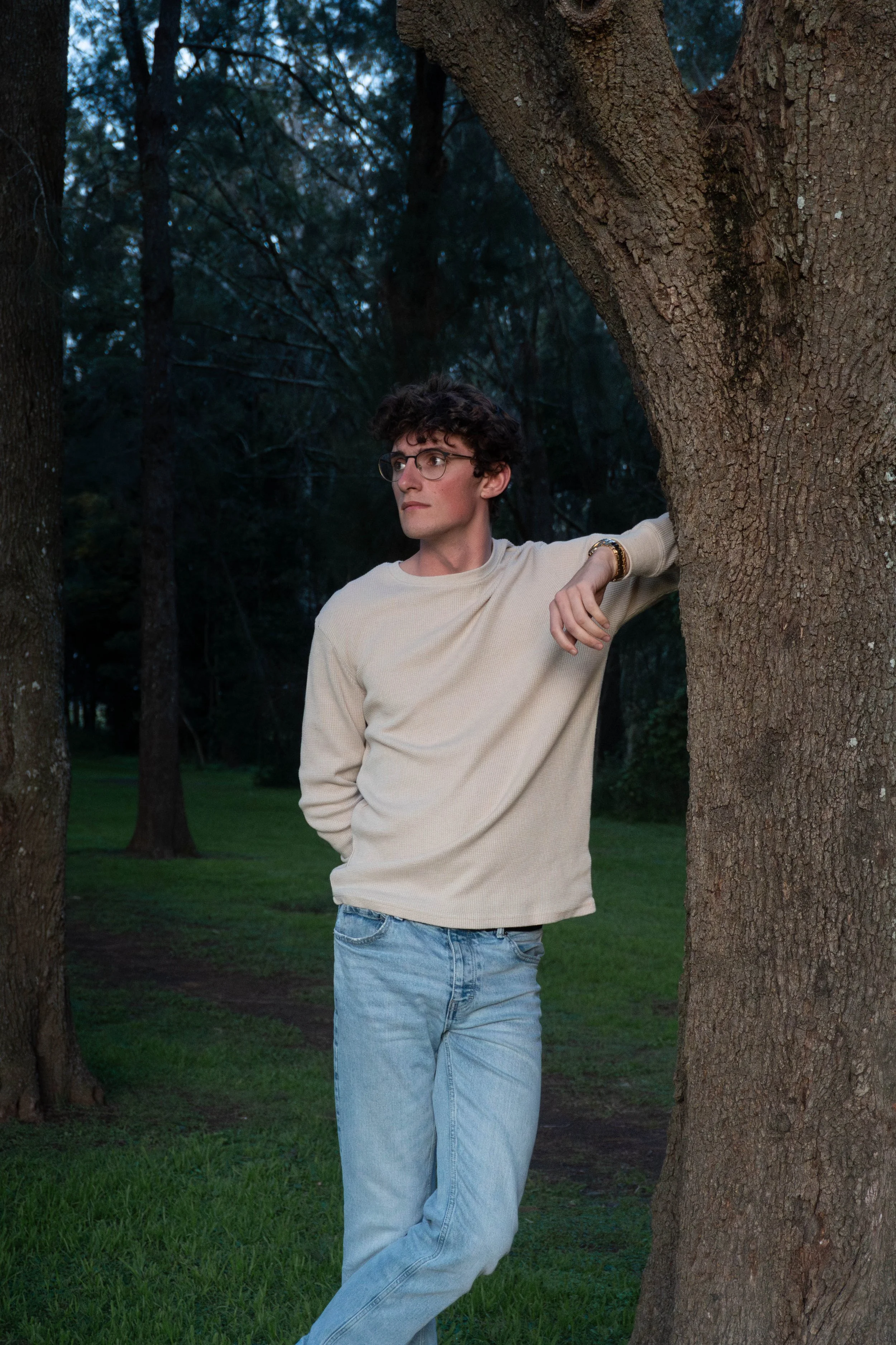 A young man with curly dark hair, glasses, wearing a beige sweater and light blue jeans, leaning against a large tree outdoors during dusk, with a background of trees and green grass.