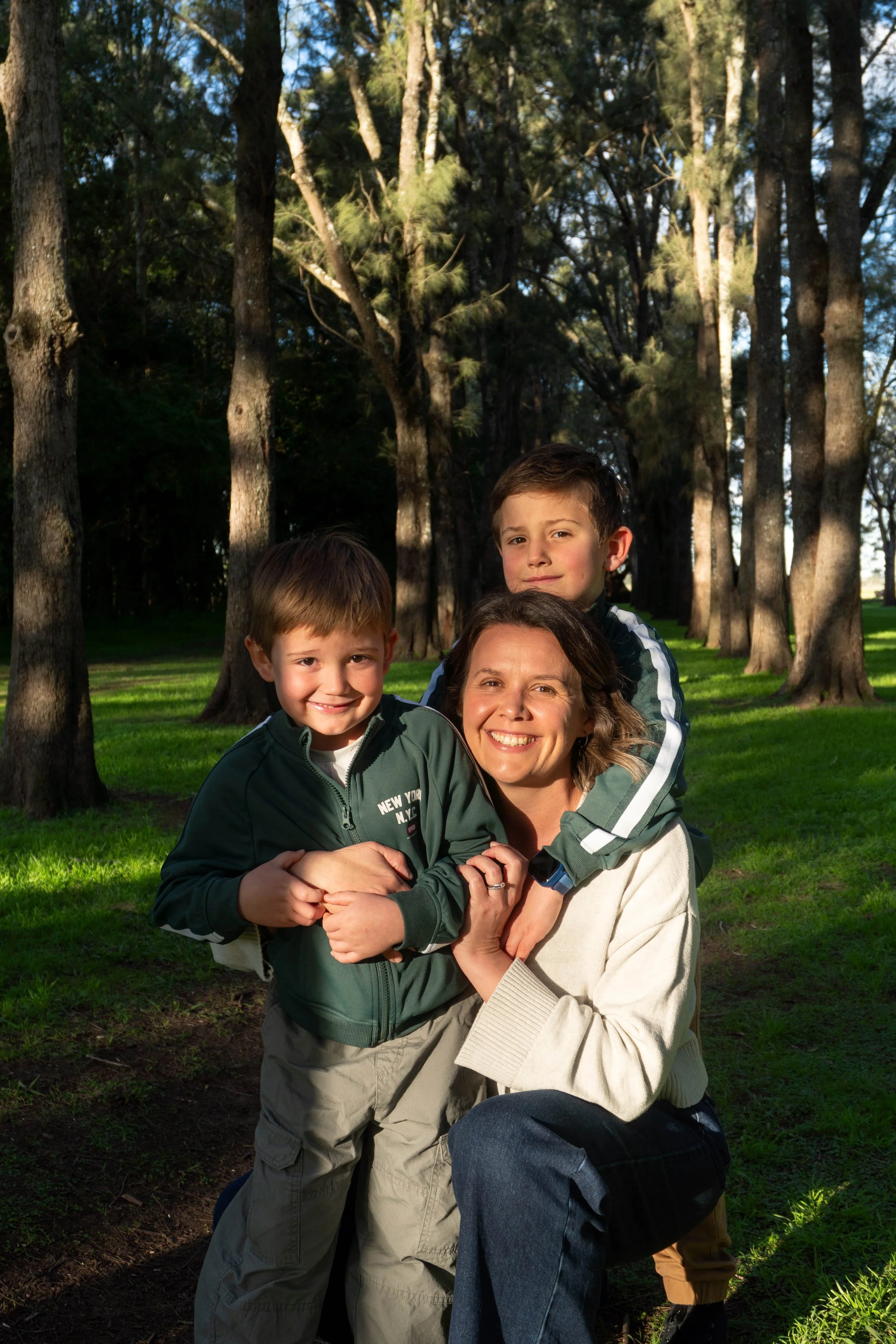 A mum with her two young boys in a forested park, smiling and hugging. The boy on the left has short brown hair, and the boy on the right has slightly longer hair. Sunlight filters through tall trees, creating a peaceful outdoor scene.