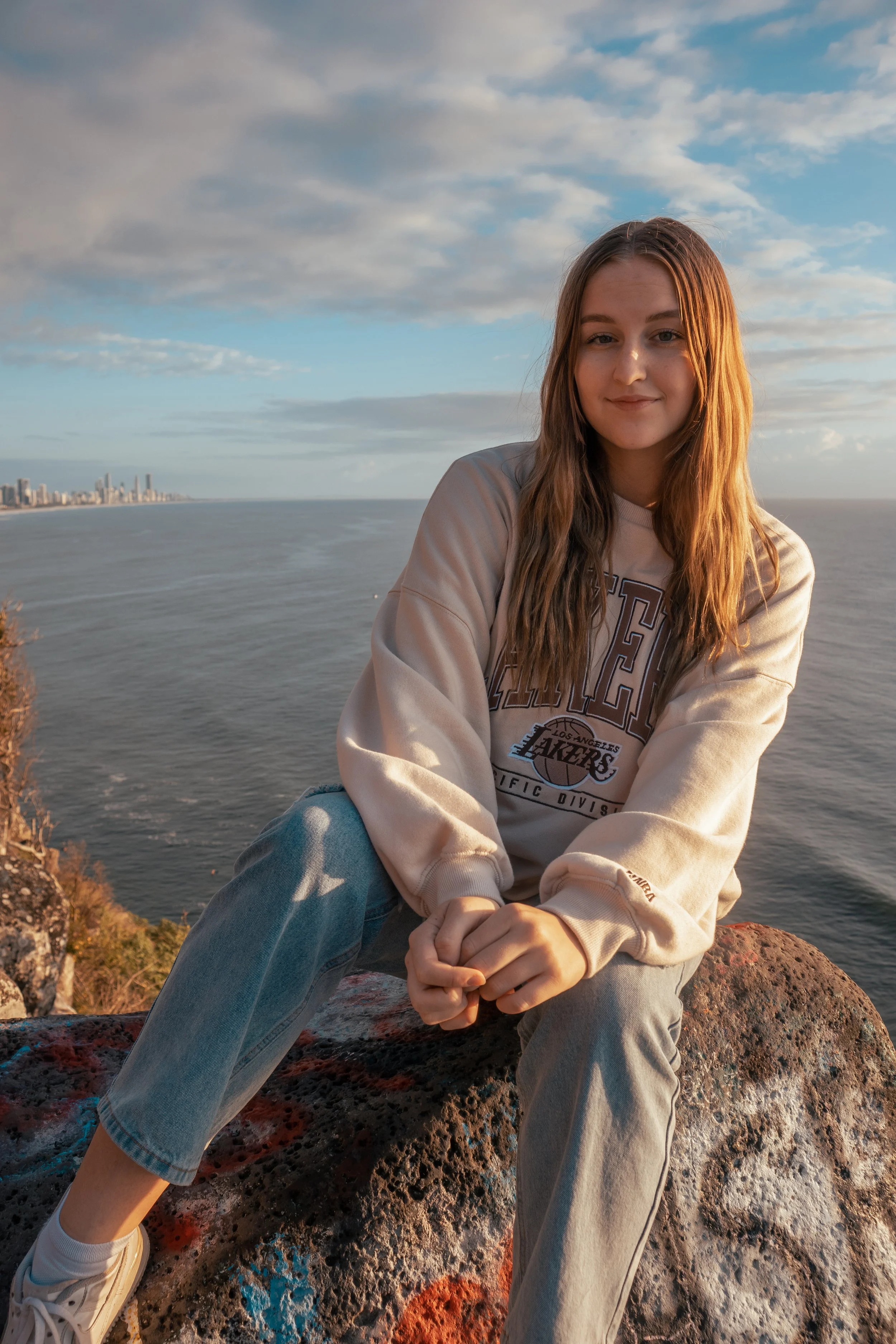 A young woman with long wavy hair, wearing a beige oversized sweatshirt with the Los Angeles Lakers logo, sitting on a rocky coast with the ocean and city skyline in the background during sunset, smiling gently at the camera.