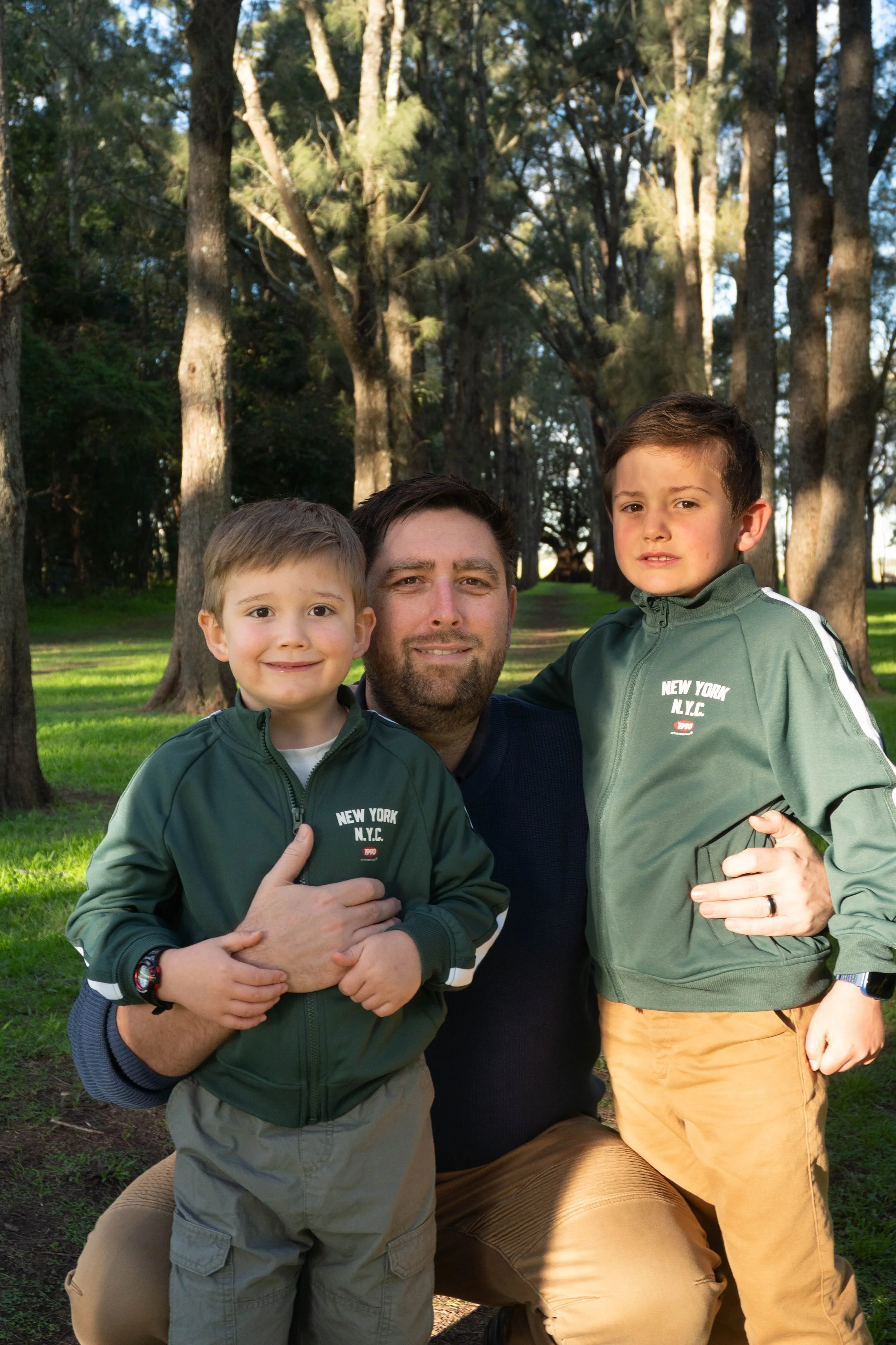 A dad with his two young boys outdoors in a park with tall trees and green grass, all wearing matching dark green jackets.