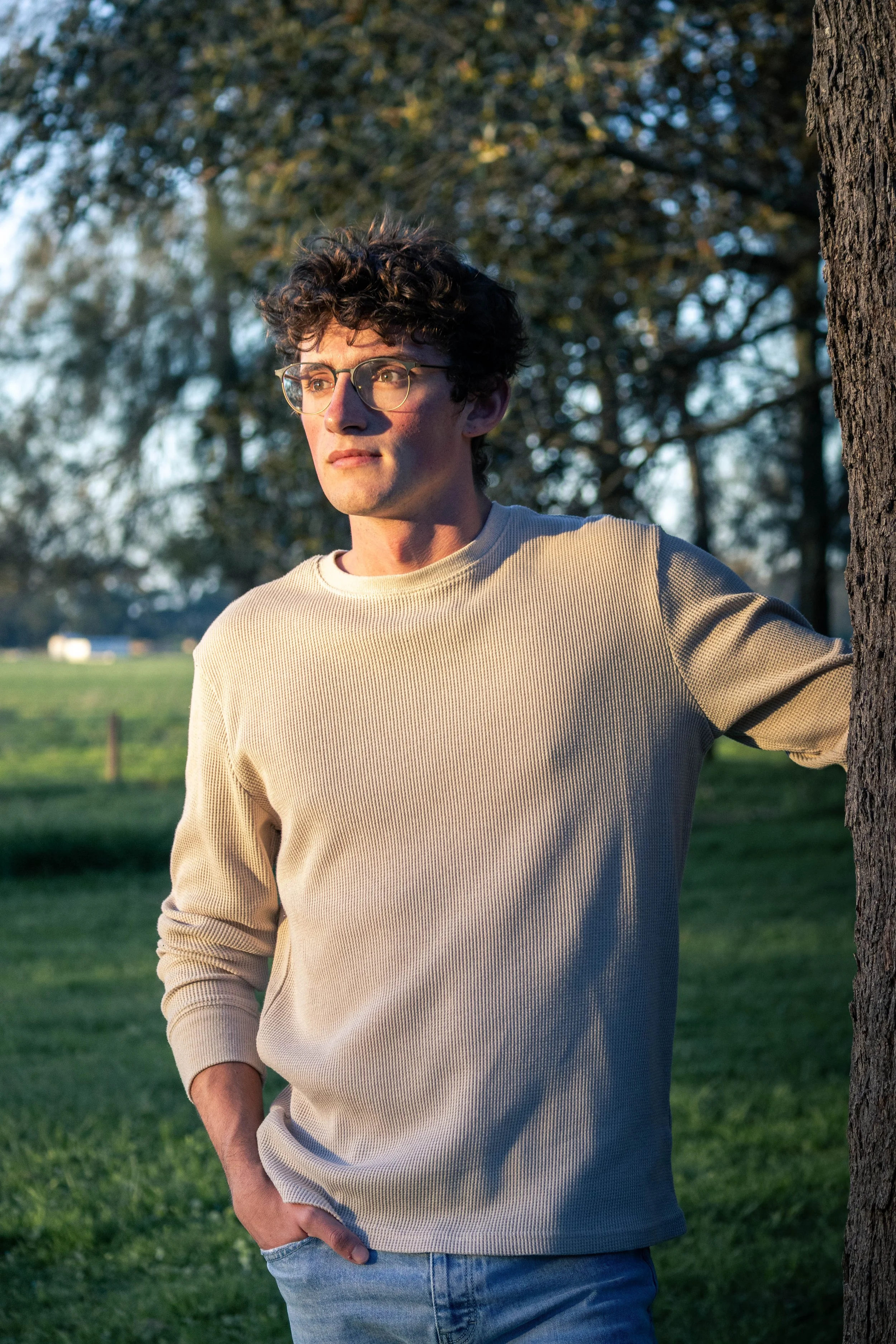 A young man with curly brown hair and glasses stands outdoors next to a tree, with a grassy field and trees in the background, sunset lighting on his face.