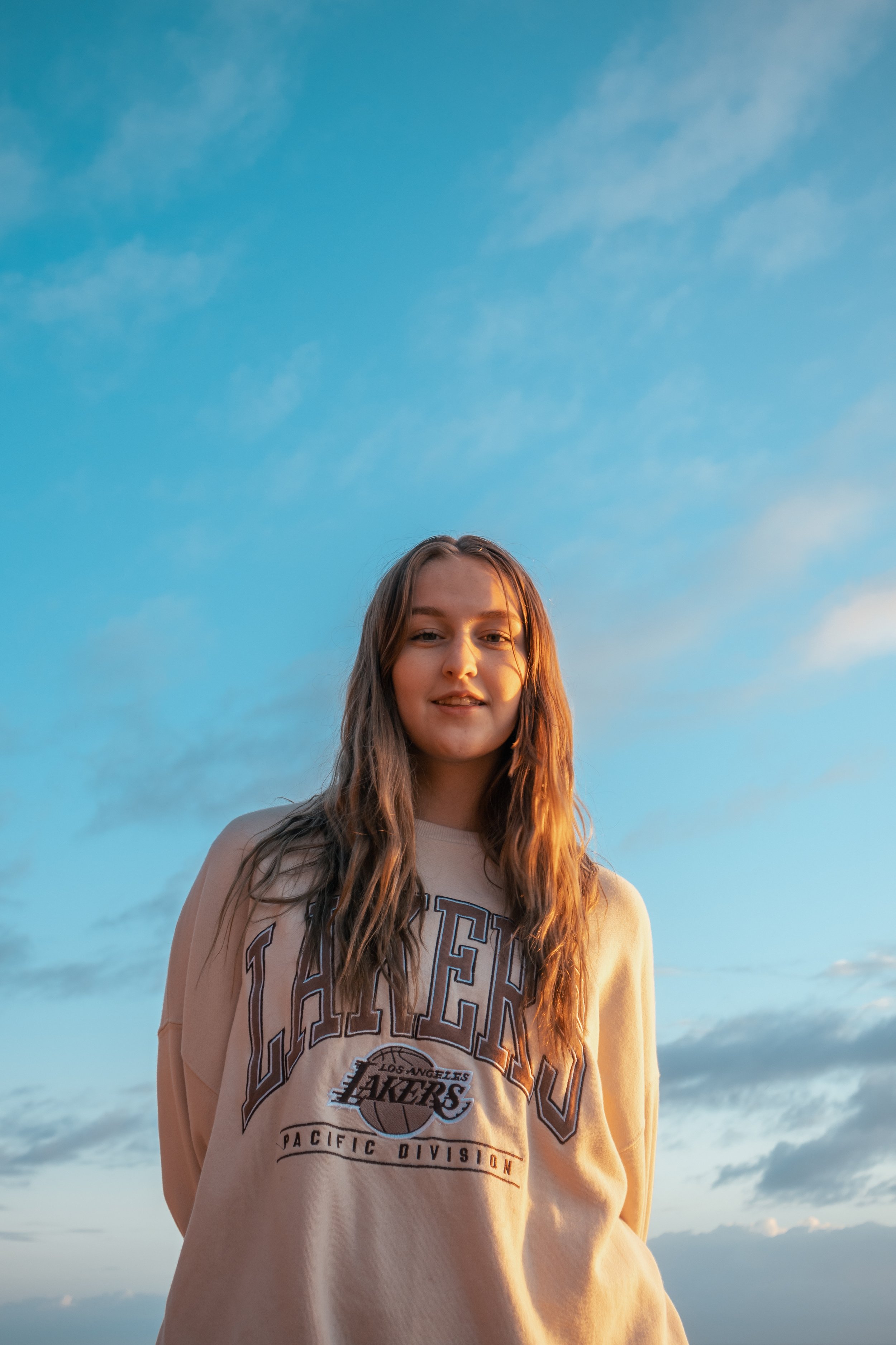 Young woman with long hair wearing a beige Los Angeles Lakers sweatshirt standing outdoors under a blue sky with scattered clouds.