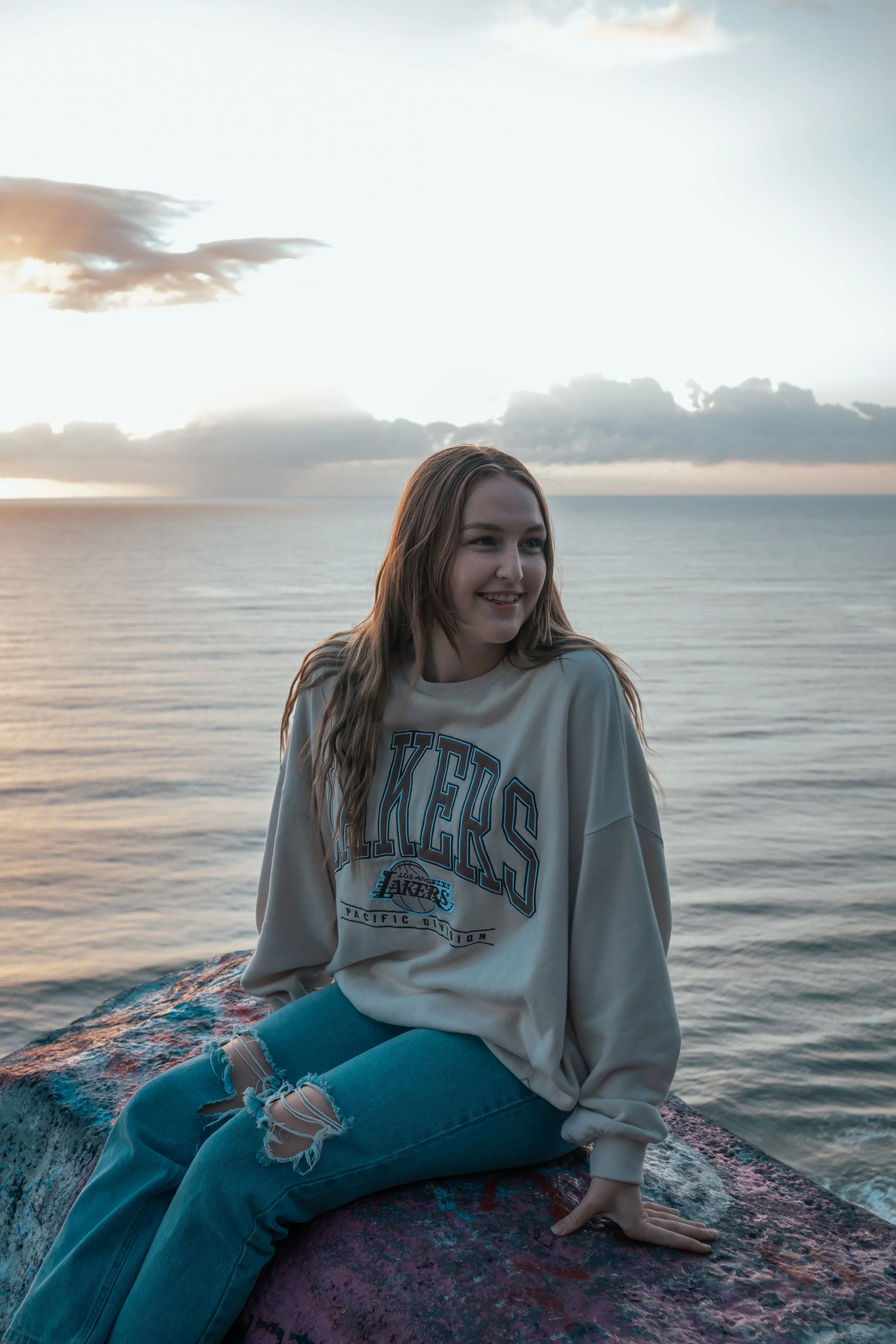 A young woman with long blonde hair sitting on a rock by the ocean during sunrise, wearing a beige Los Angeles Lakers sweatshirt.