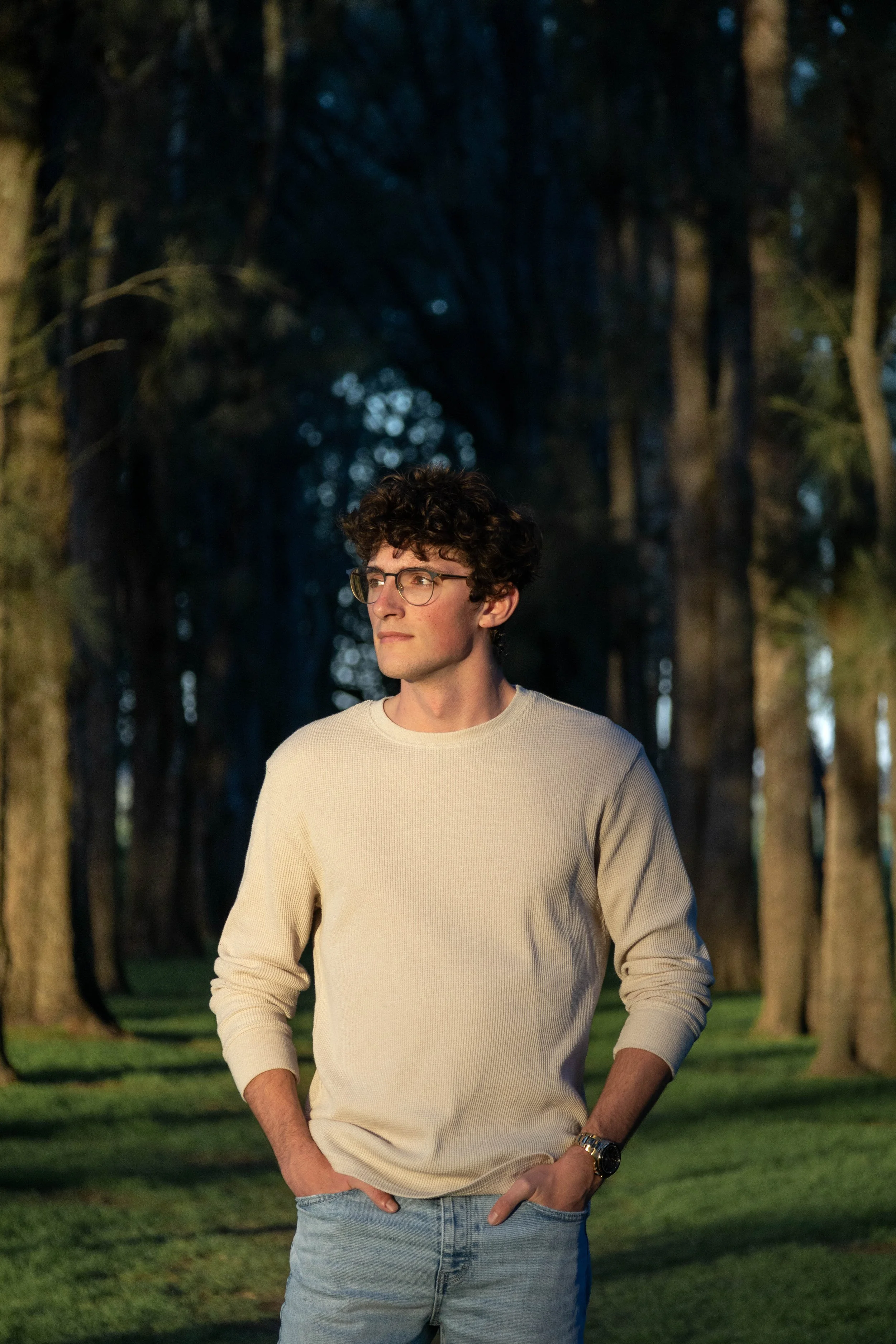 A young man with curly brown hair, glasses, and light skin, standing outdoors in front of tall trees during early evening, wearing a cream-colored sweater, light blue jeans, and a gold watch, with his hands in his pockets and a serious expression.