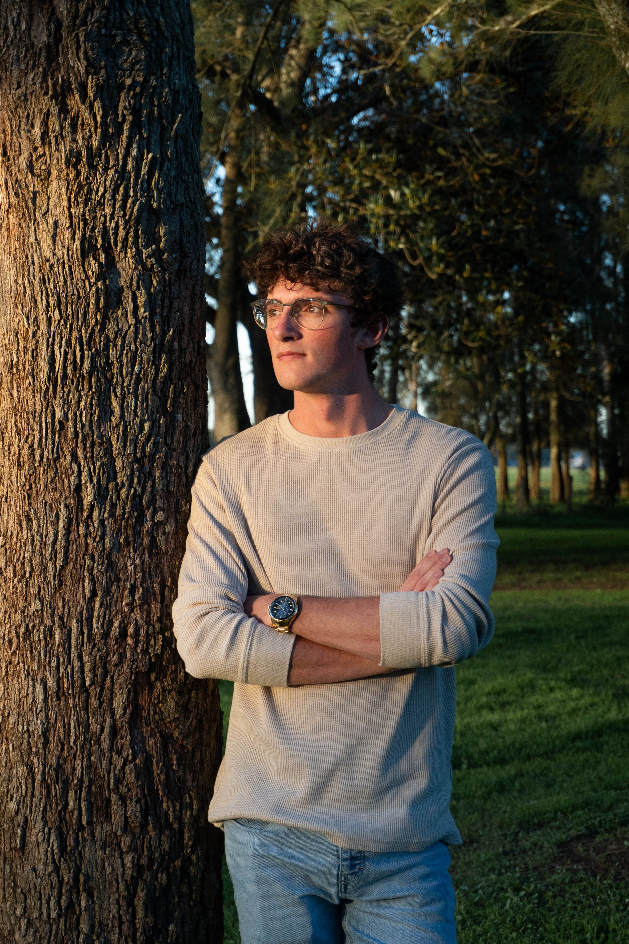 A young man with curly hair and glasses standing outdoors next to a large tree trunk, with arms crossed, during sunset in a park with greenery and other trees in the background.