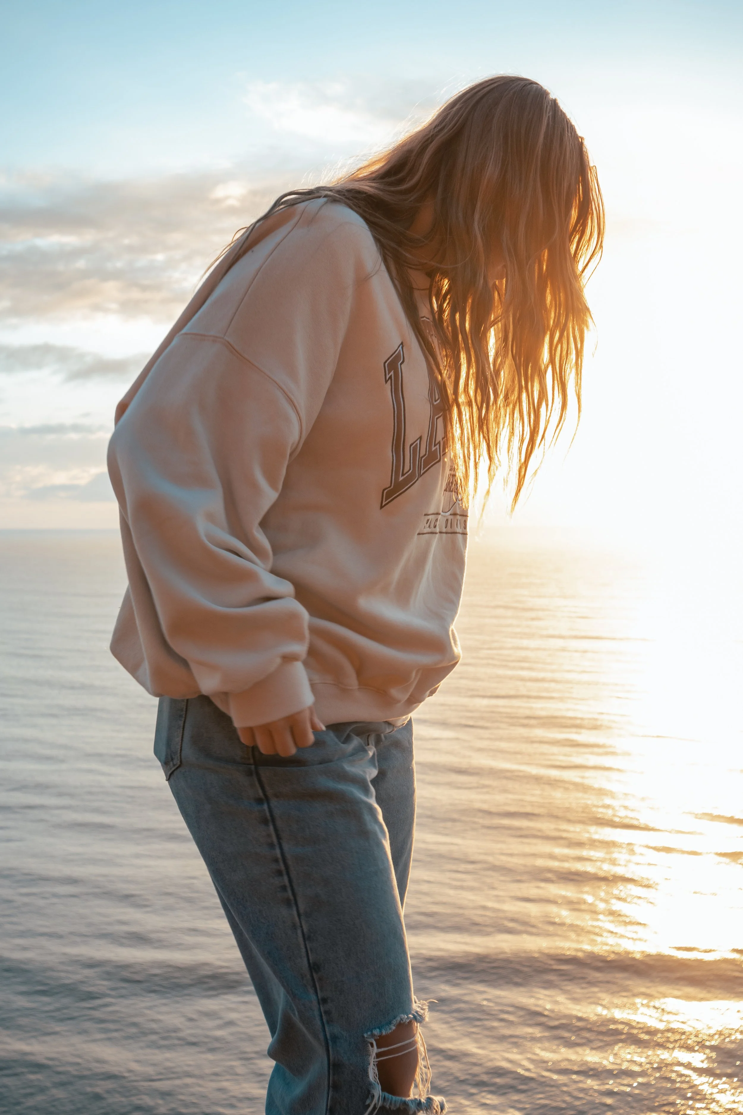 A woman with long wavy hair wearing a beige sweatshirt and ripped jeans standing near water during sunrise.