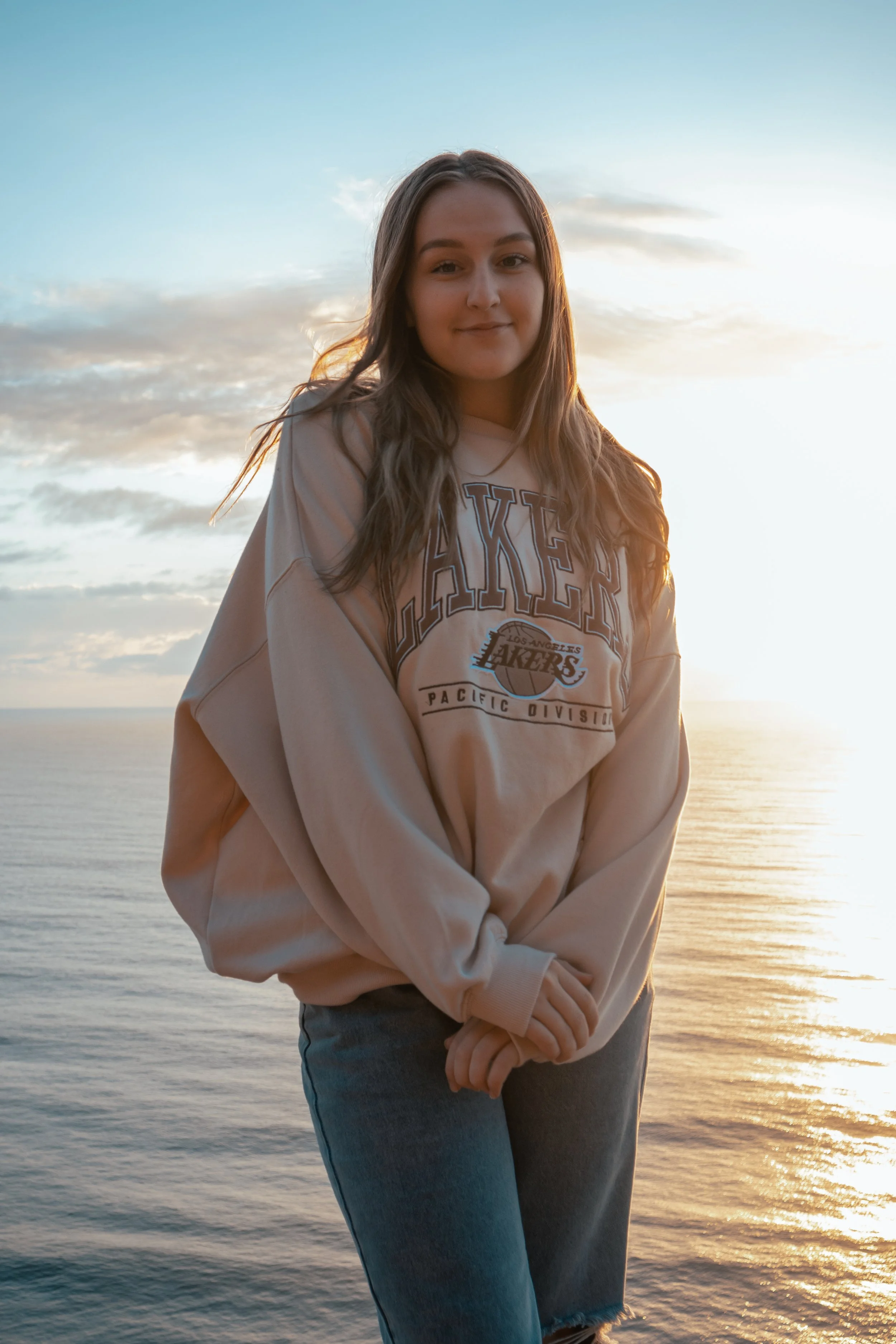 A young woman with long hair standing near the water at sunrise, wearing a beige Lakers sweatshirt and blue jeans.
