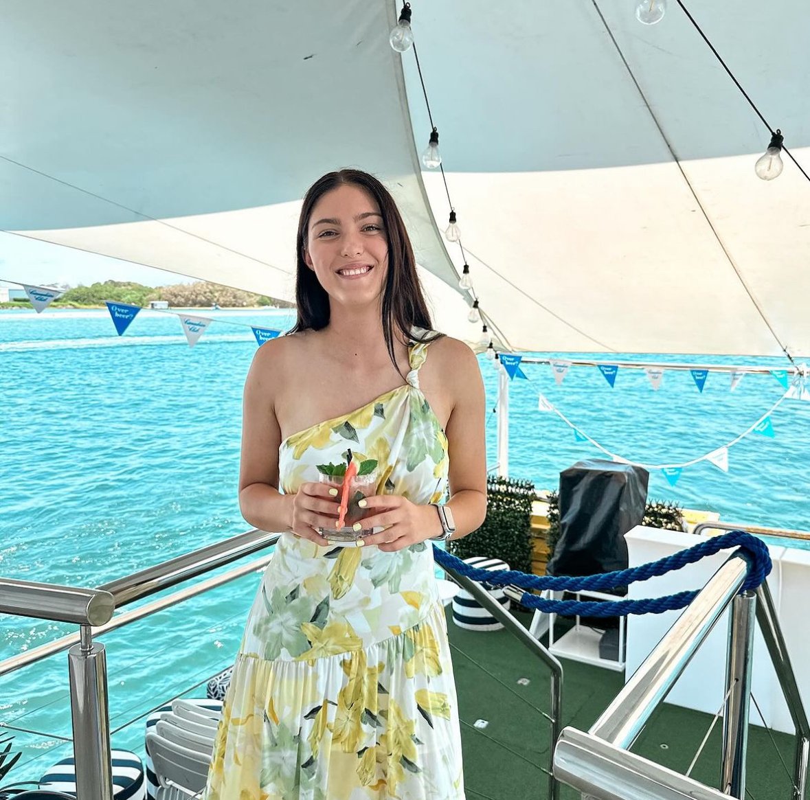 A young woman in a yellow and white floral dress holding a drink on a boat with ocean water in the background.