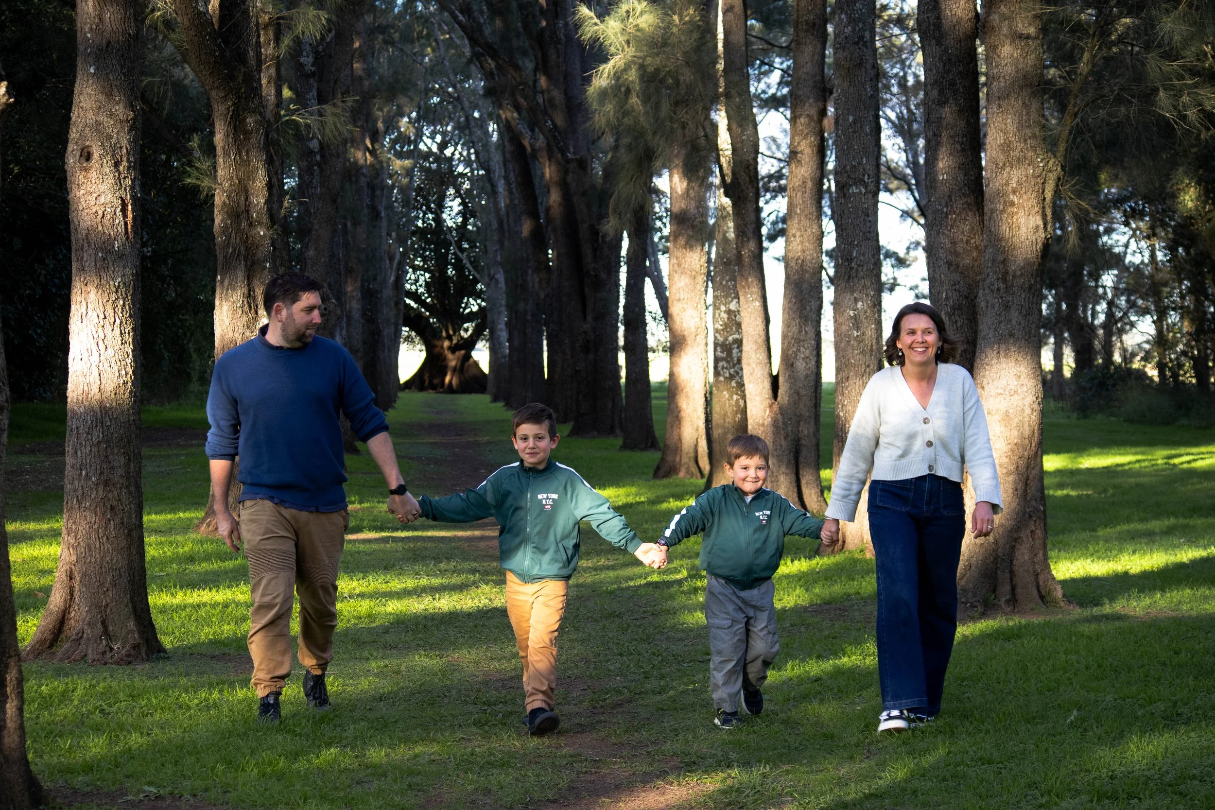 A family of four walking through a forest, holding hands, with the adults smiling and the children looking ahead.