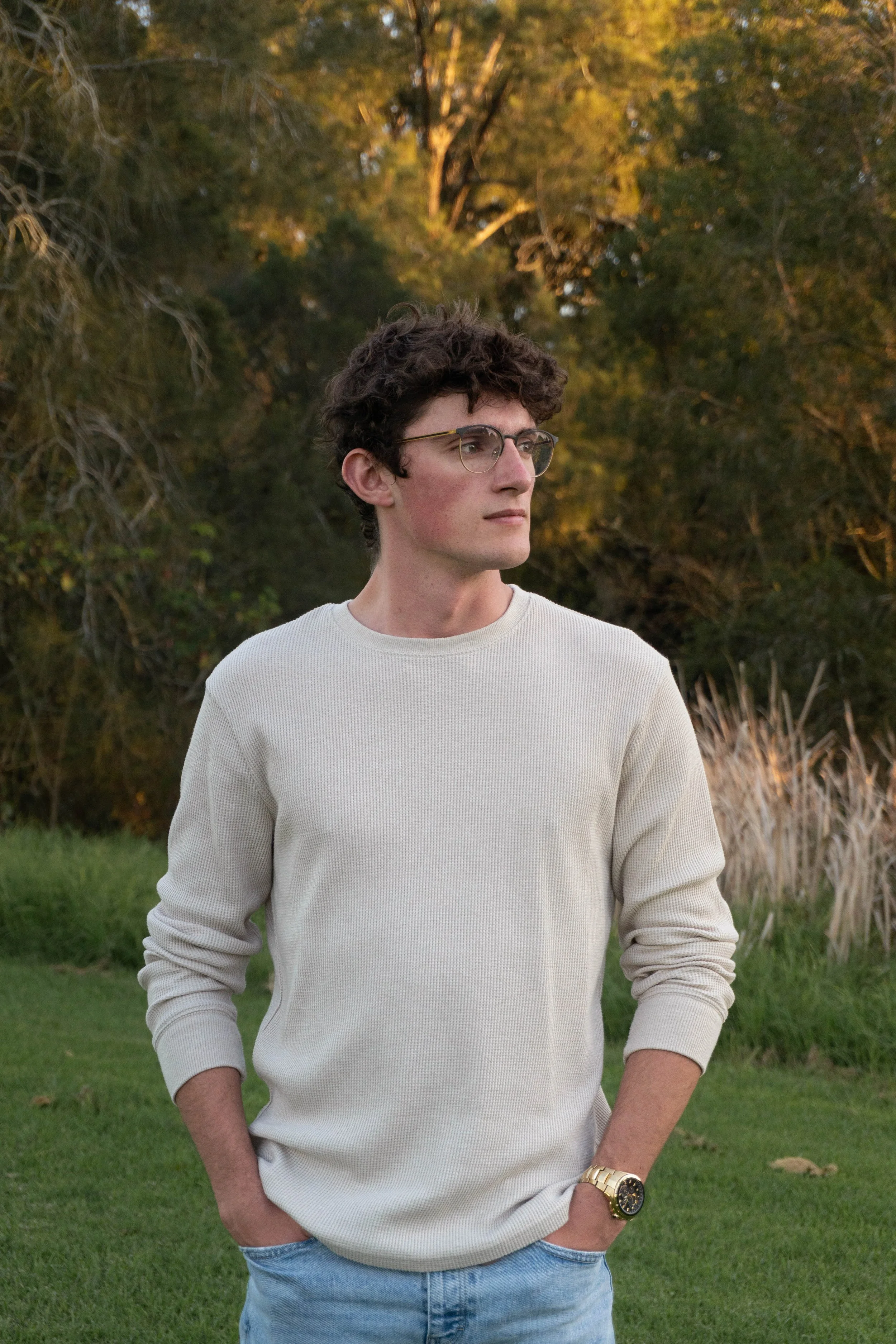 A young man with curly dark hair and glasses wearing a cream sweater and blue jeans standing outdoors with hands in pockets, surrounded by green grass and trees in the background.