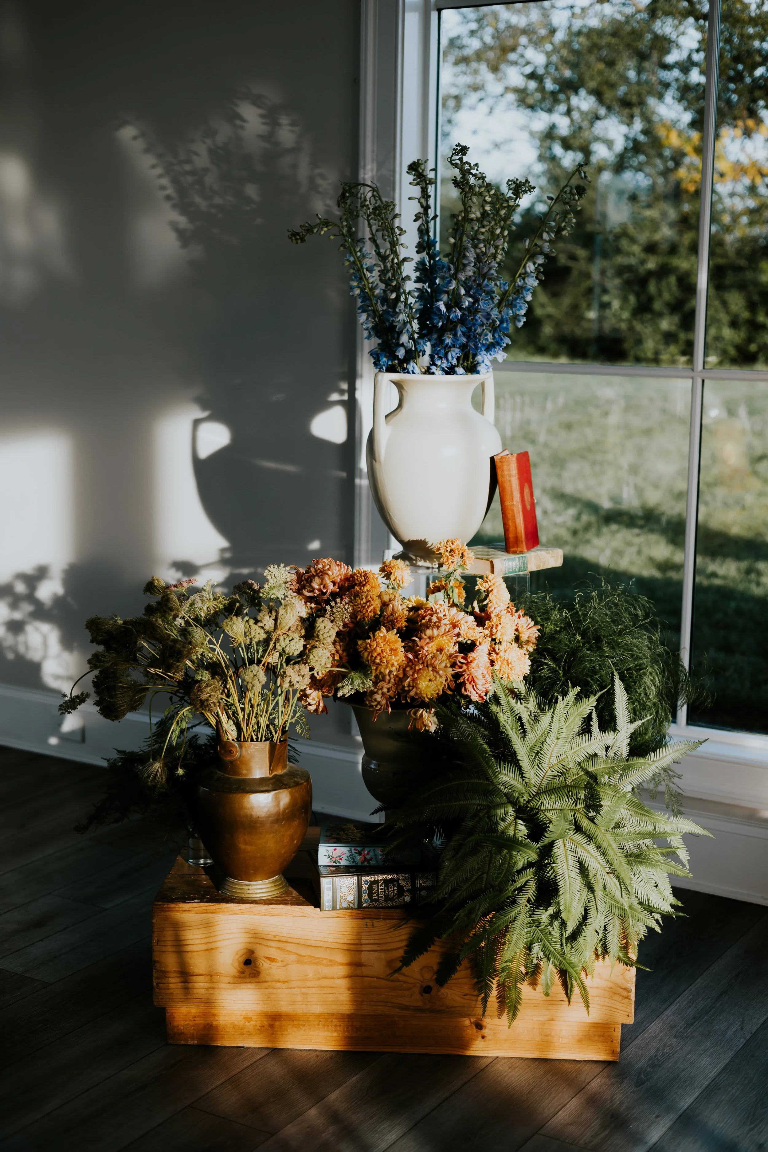 A wooden planter box filled with various flowers and ferns, placed near a window with natural light. On top of the box, a white ceramic vase with tall blue and green flowers is positioned beside a stack of books.