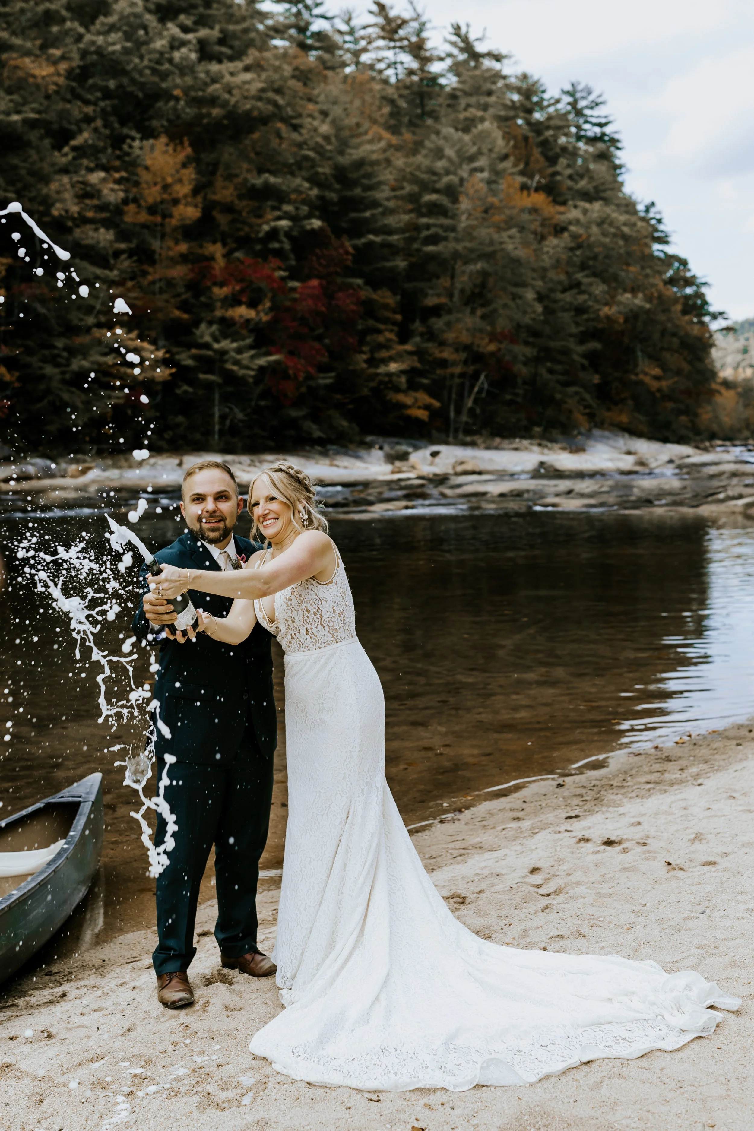 A newlywed couple celebrating by a riverbank, with the bride in a white lace wedding dress and the groom in a dark suit, as champagne sprays around them during daytime with trees in fall foliage in the background.