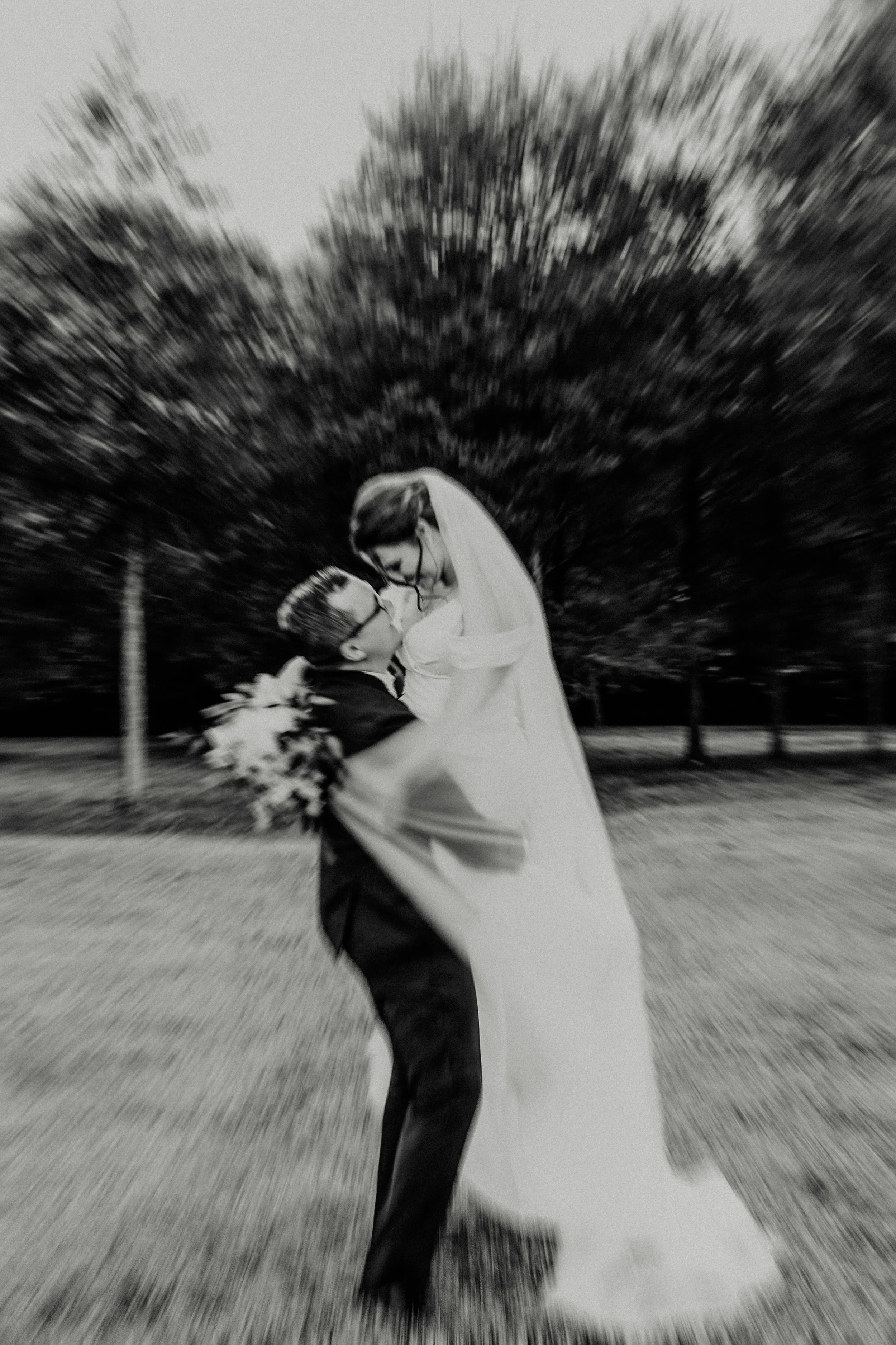 Black and white photo of a person in a graduation cap and gown, holding a bouquet, embracing another person, with a blurred background of trees and a pathway.