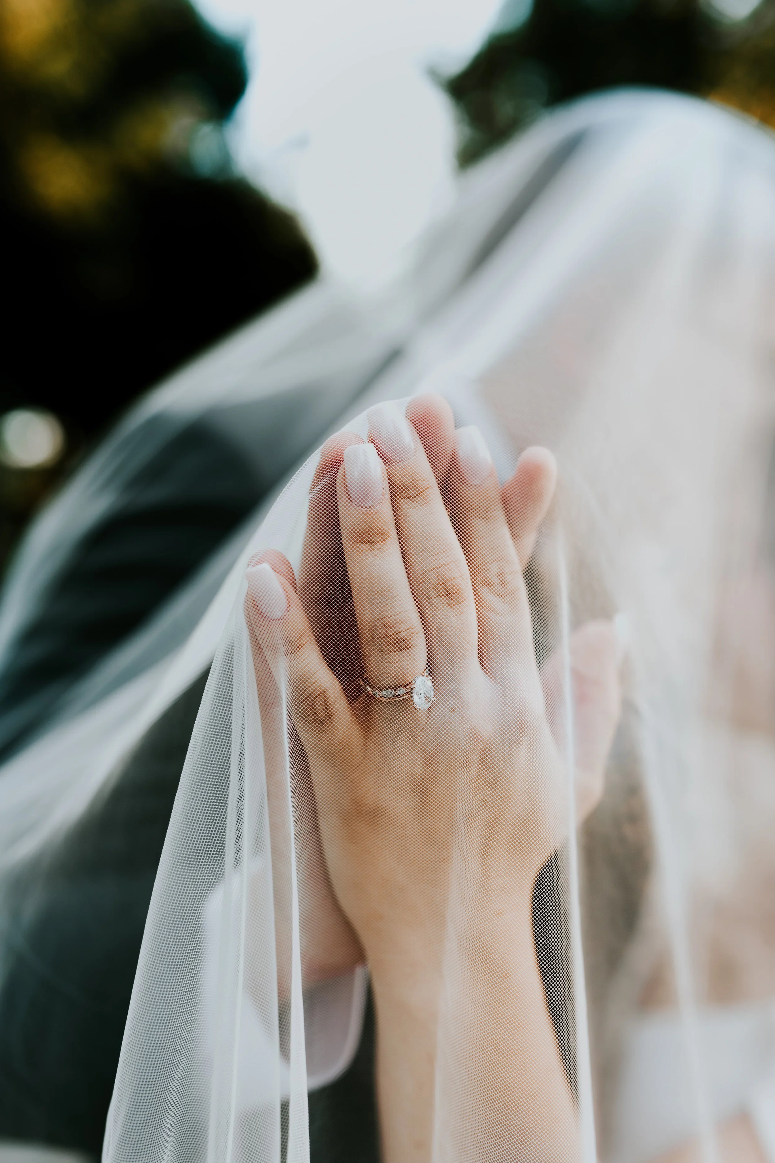 Close-up of a bride's hand with a diamond ring, touching a car window with a sheer veil in the background.