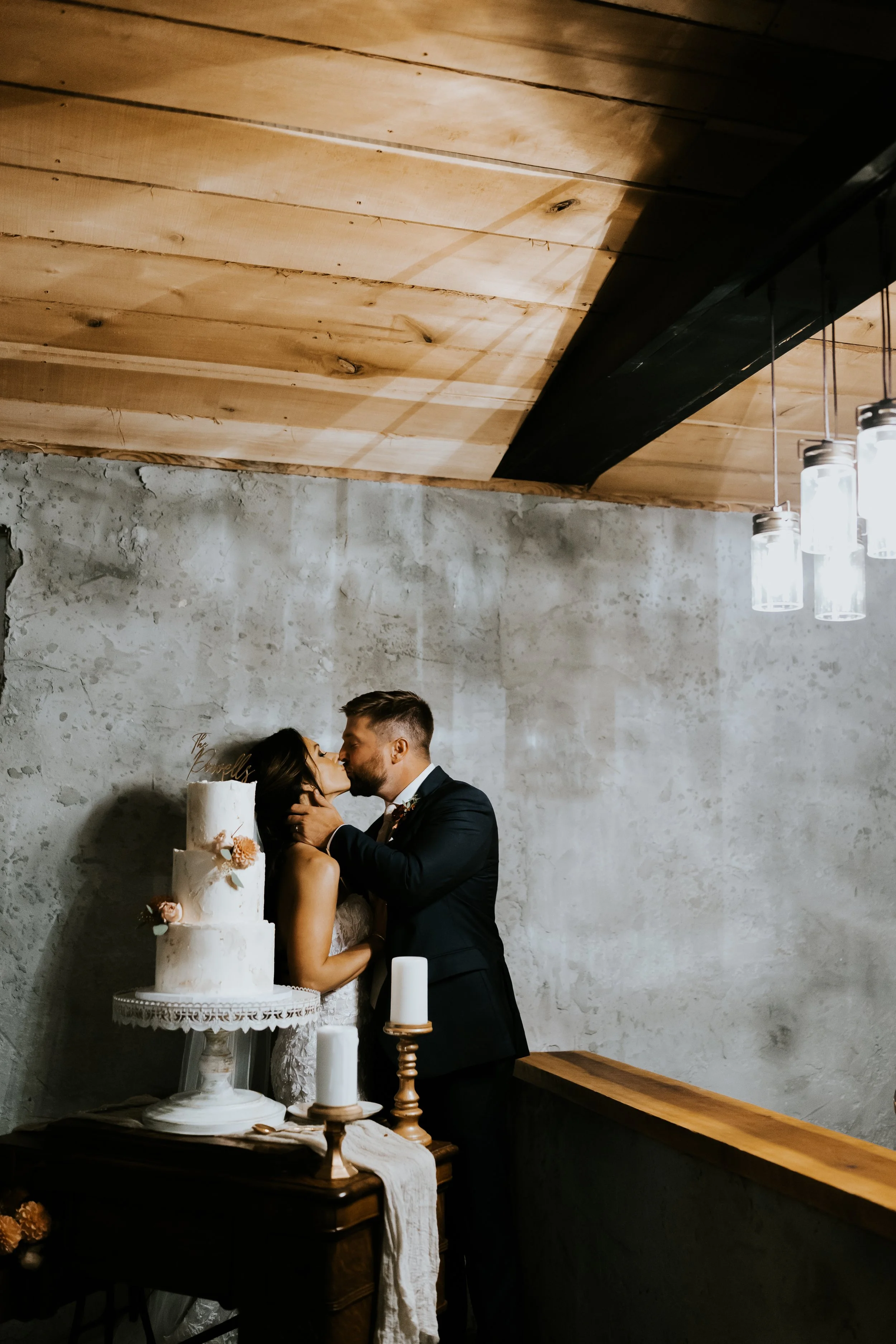 A couple sharing a kiss during their wedding reception, with a wedding cake, candles, and decorative items on a table in front of them, in an industrial-style room with a concrete wall and wooden ceiling.