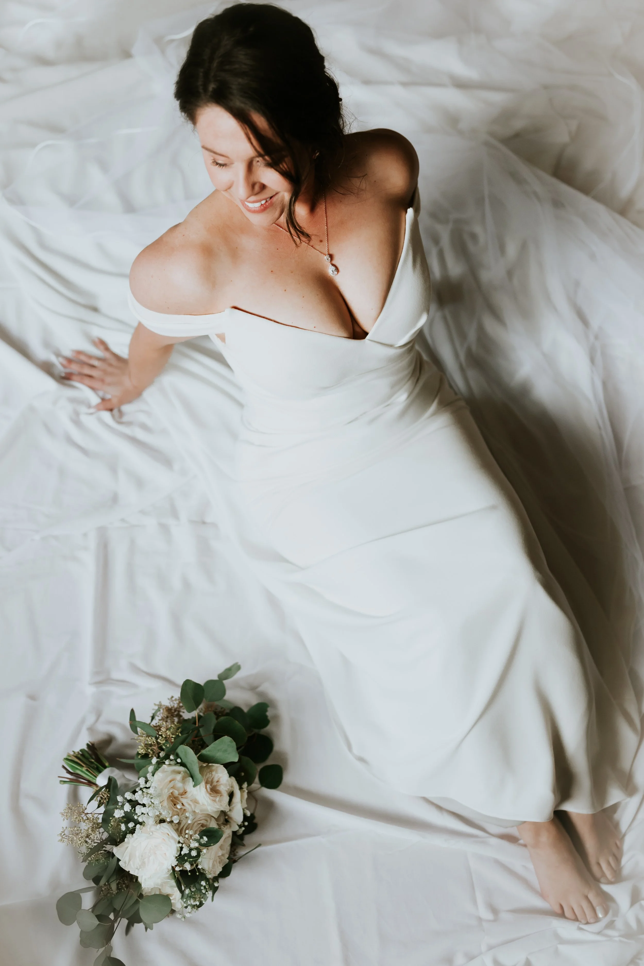 A woman with dark hair wearing a strapless white dress and a necklace, sitting on a bed with white sheets, while looking down and smiling. A bouquet of white flowers and green leaves rests beside her on the bed.