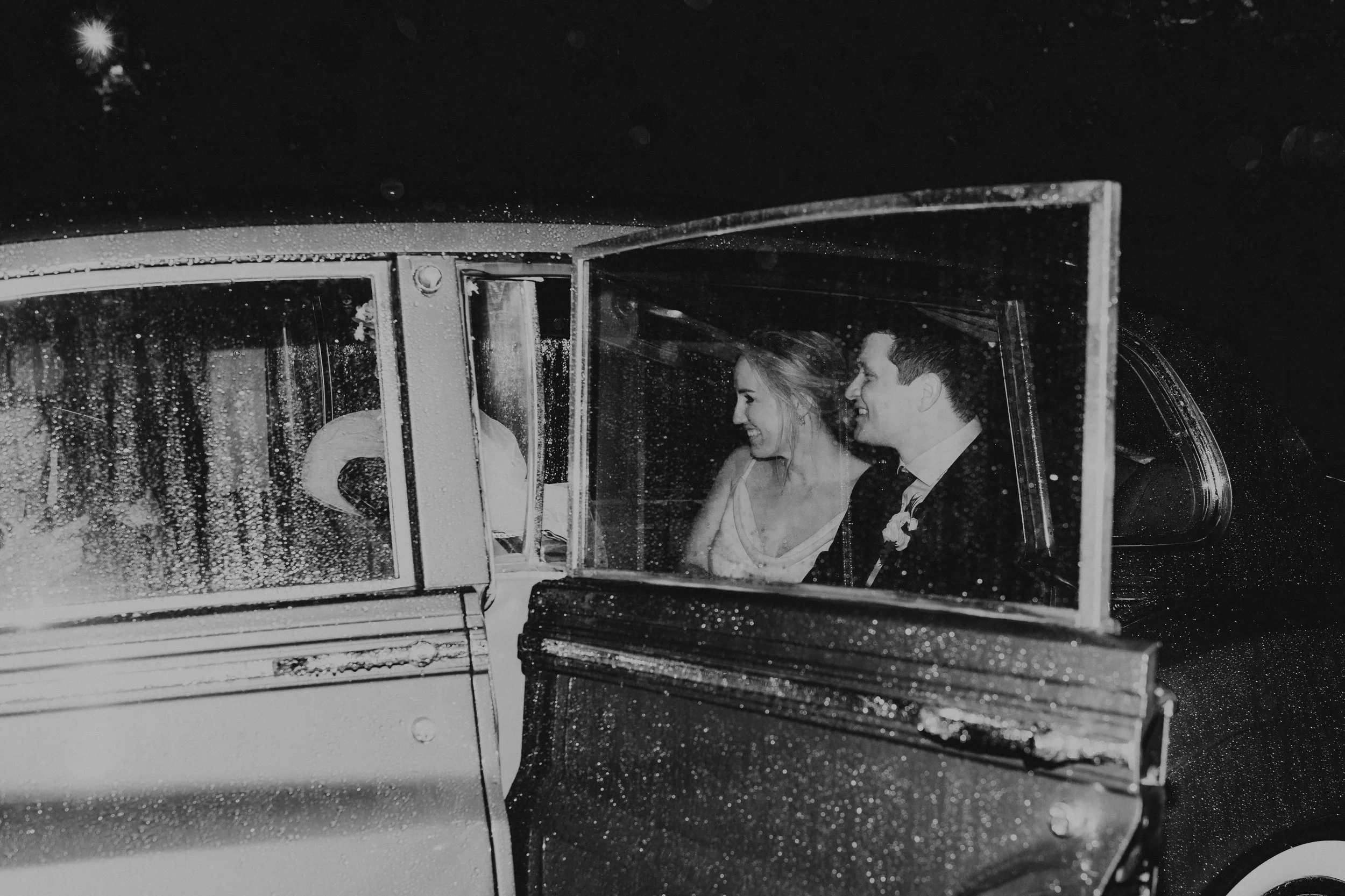 A black and white photo of a woman and a man inside a vintage car, with rain on the windows and at night, possibly during a rainy night.
