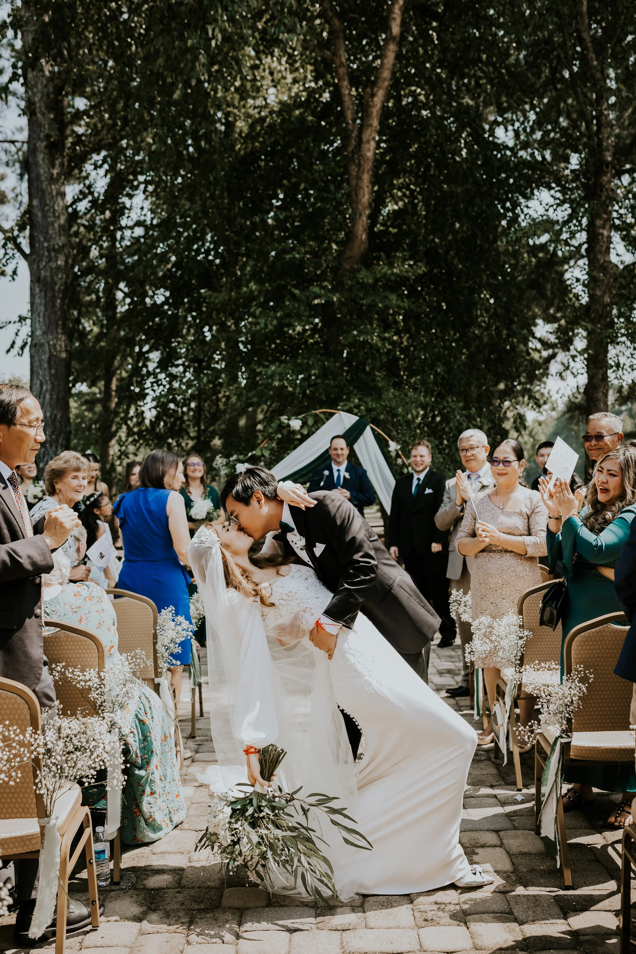 A bride and groom kissing during an outdoor wedding ceremony, surrounded by seated guests and onlookers, under a large tree with a tent in the background.