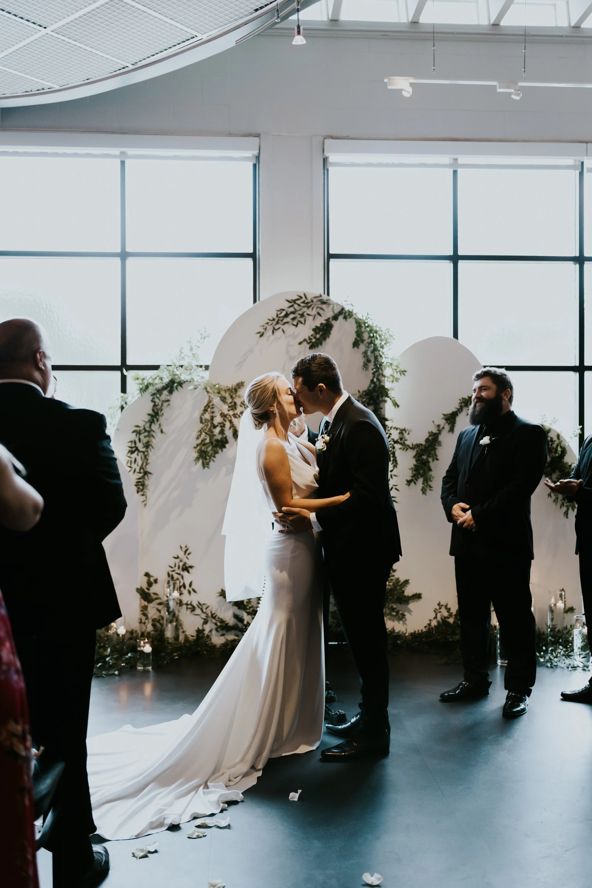 A couple in wedding attire sharing a kiss during their wedding ceremony, standing in front of a floral backdrop, surrounded by friends and family in a modern, well-lit indoor space.