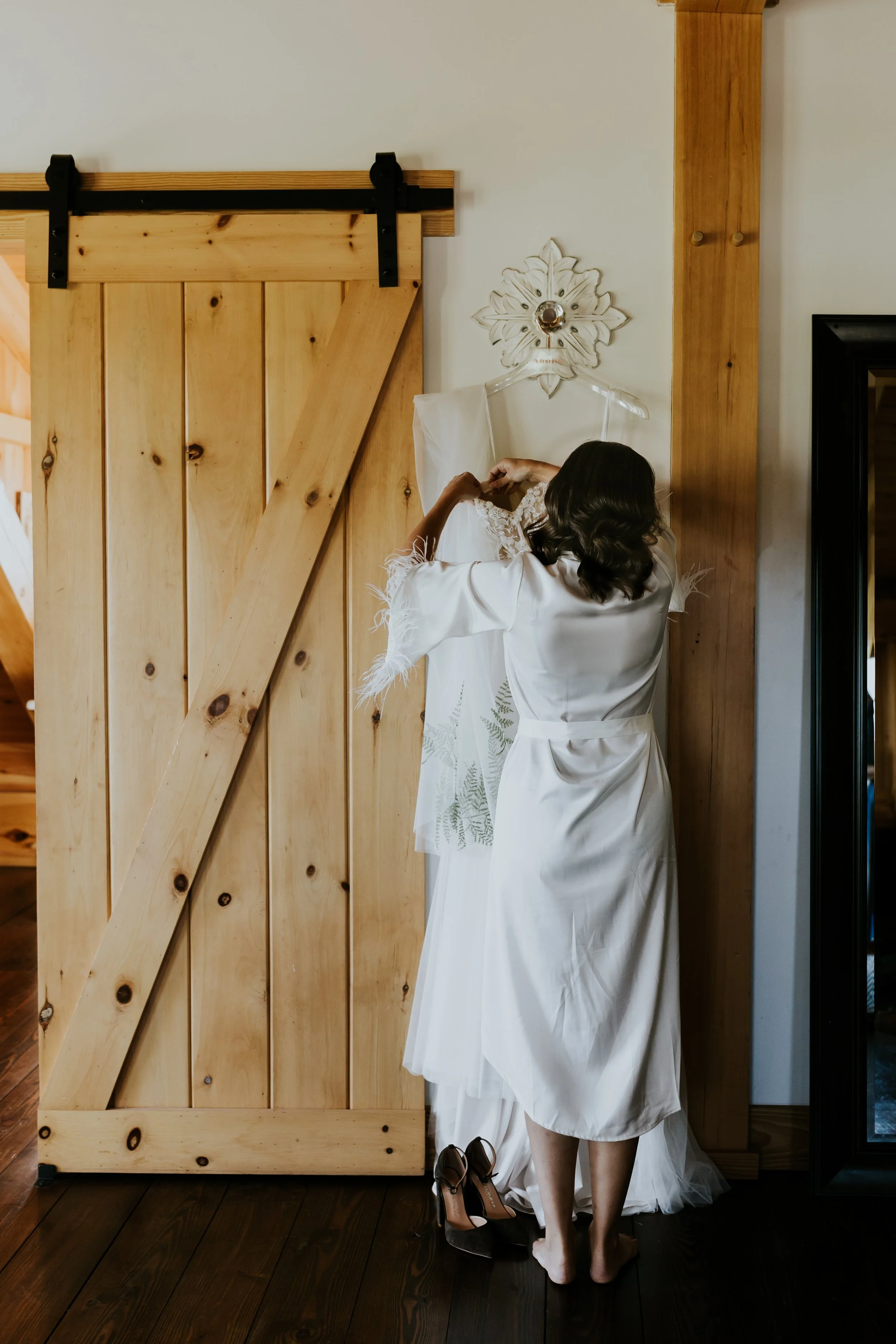 A woman with dark hair in a white satin robe is hanging a wedding dress on a wall-mounted hanger under a decorative metal wall sconce in a room with wooden accents.