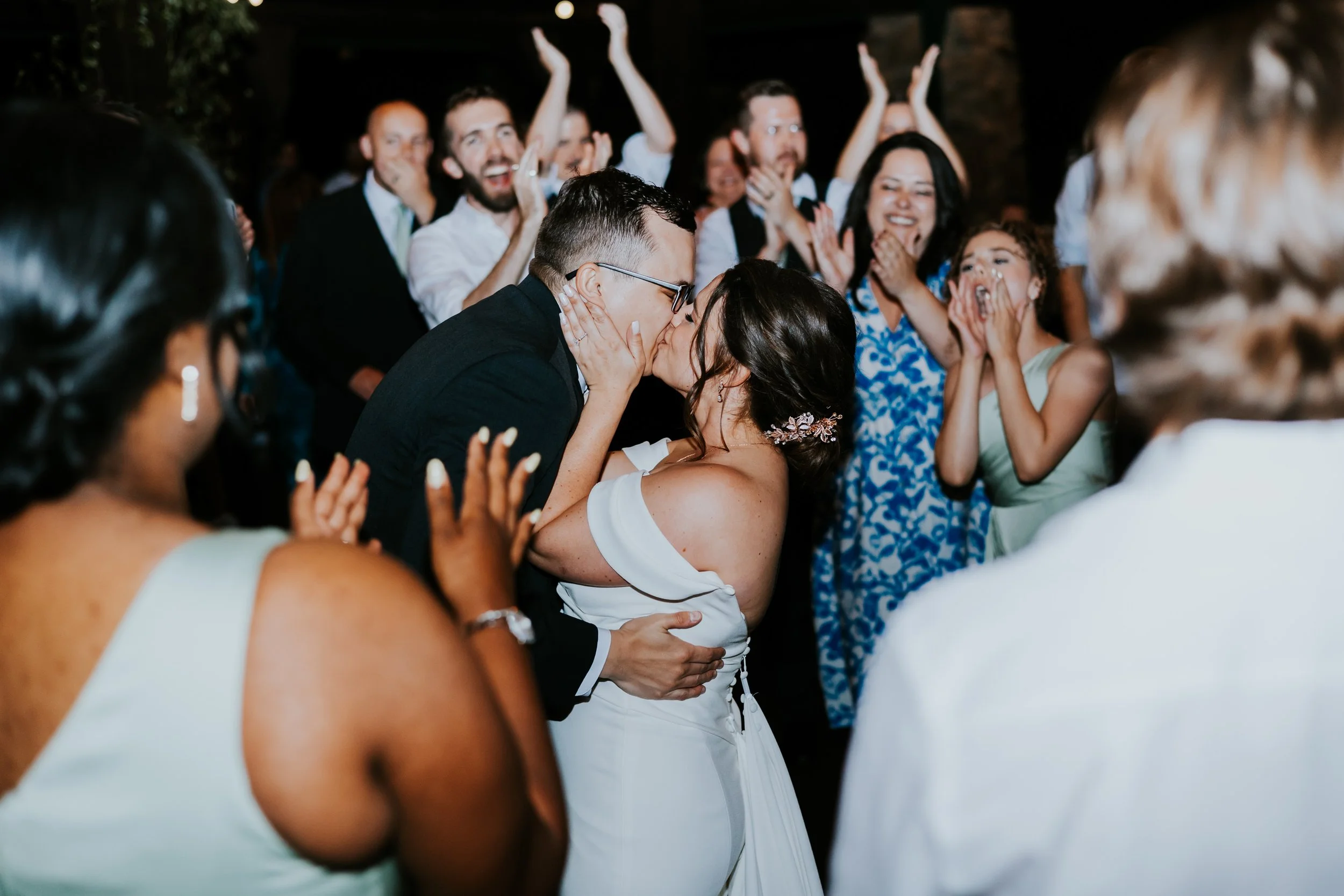 A bride and groom kiss as their guests cheer and celebrate at a wedding reception.