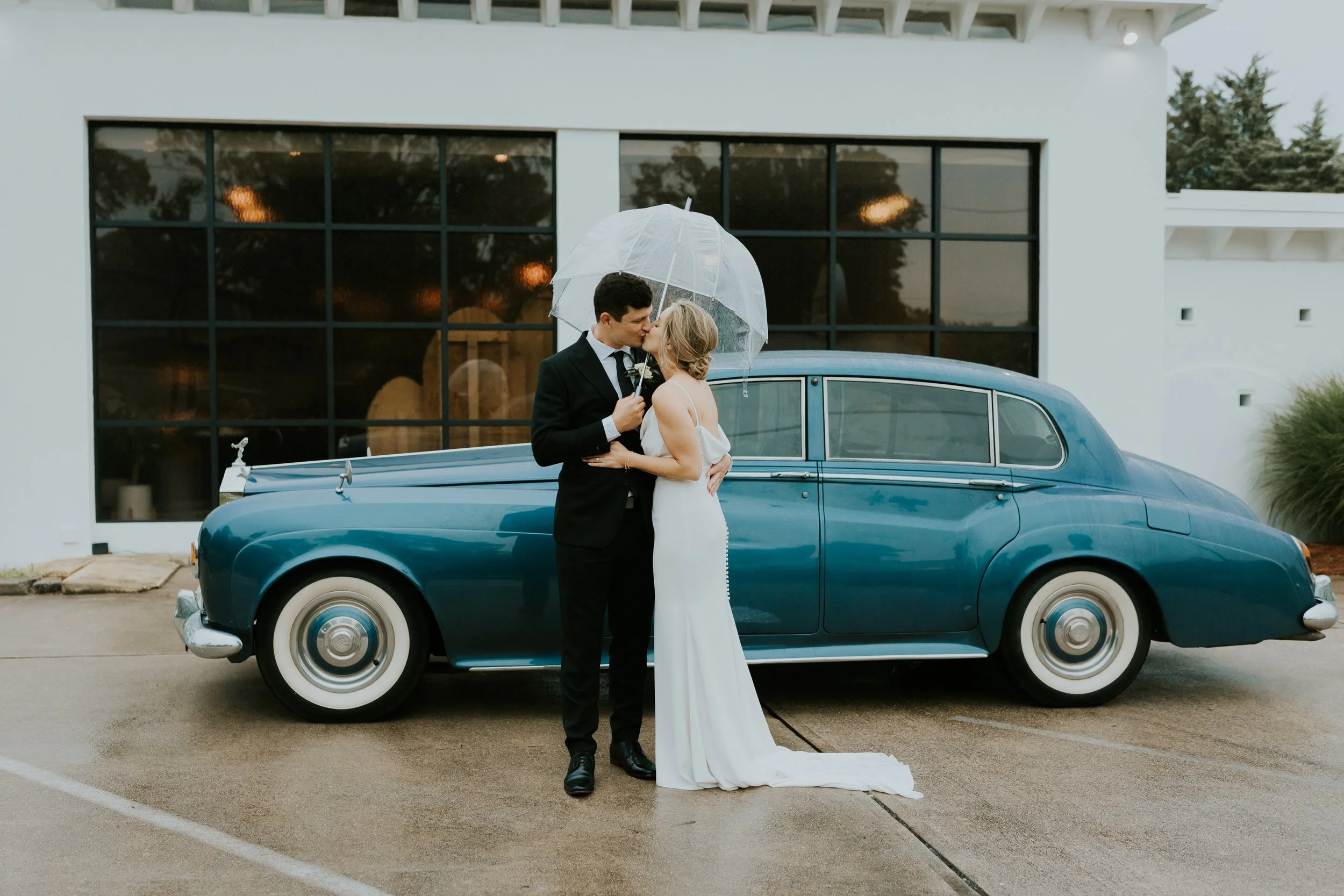 A couple dressed in wedding attire sharing a kiss under a clear umbrella in front of a vintage blue car outside a modern white building.