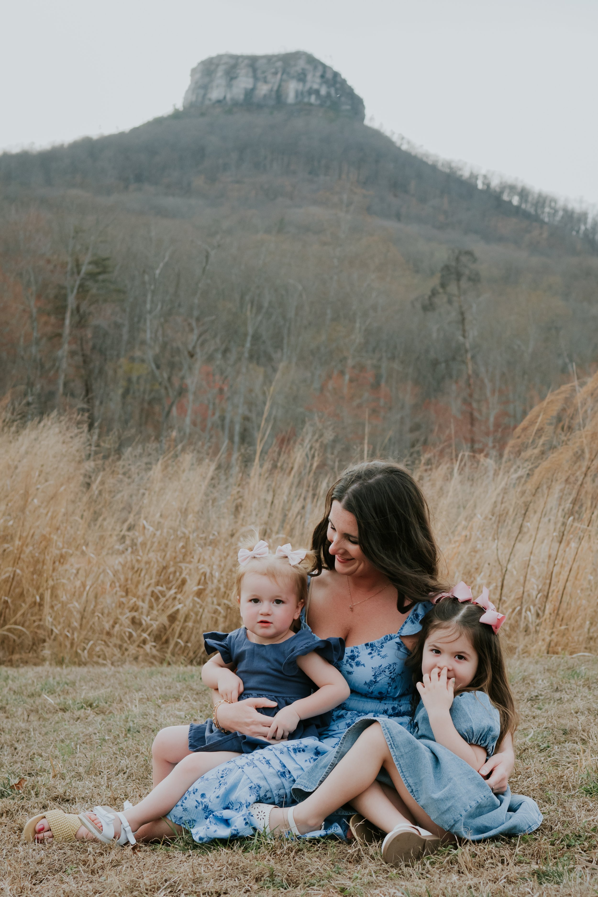 A woman with long dark hair smiling and sitting on the grass, holding two young girls, one with light hair and the other with dark hair, in an outdoor setting with tall dry grass and a mountain in the background during daytime.