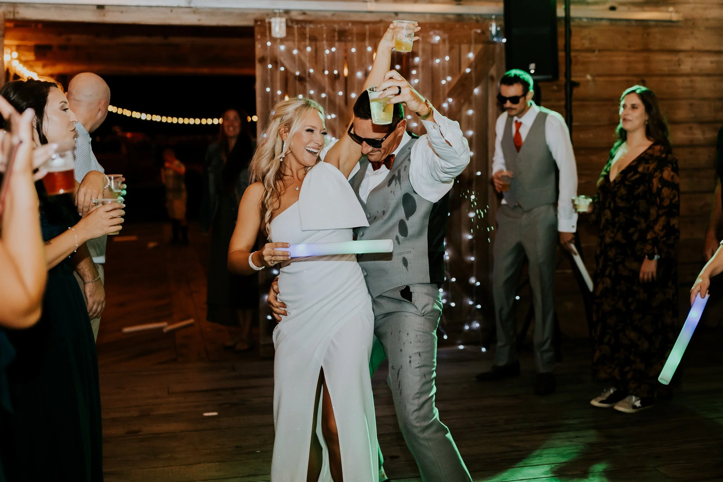 A bride and groom dancing at their wedding reception, surrounded by guests in a rustic wooden venue with string lights.