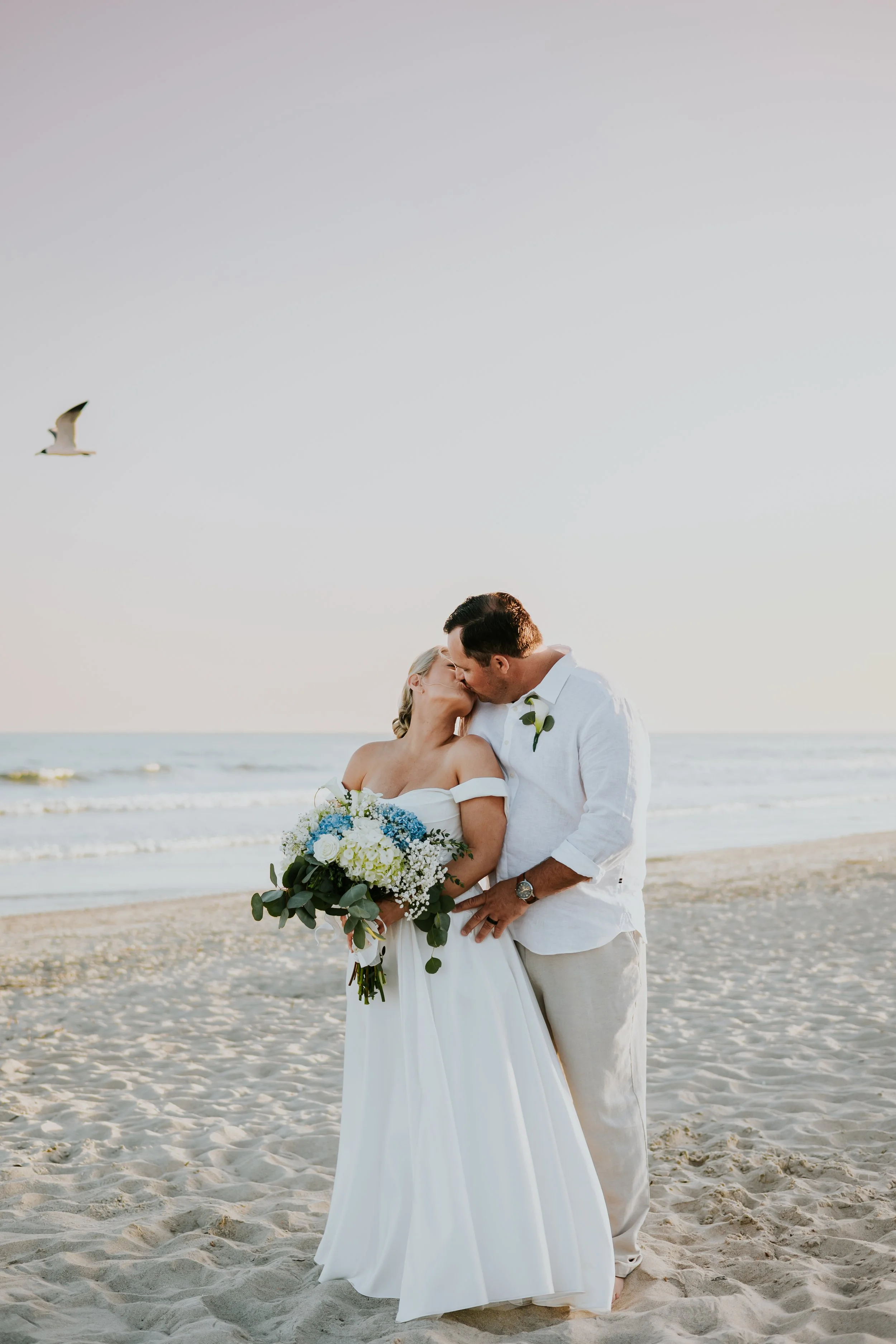 A bride and groom sharing a kiss on the beach, with the bride holding a bouquet of white, blue, and green flowers, during sunset with an ocean backdrop and a seagull flying overhead.