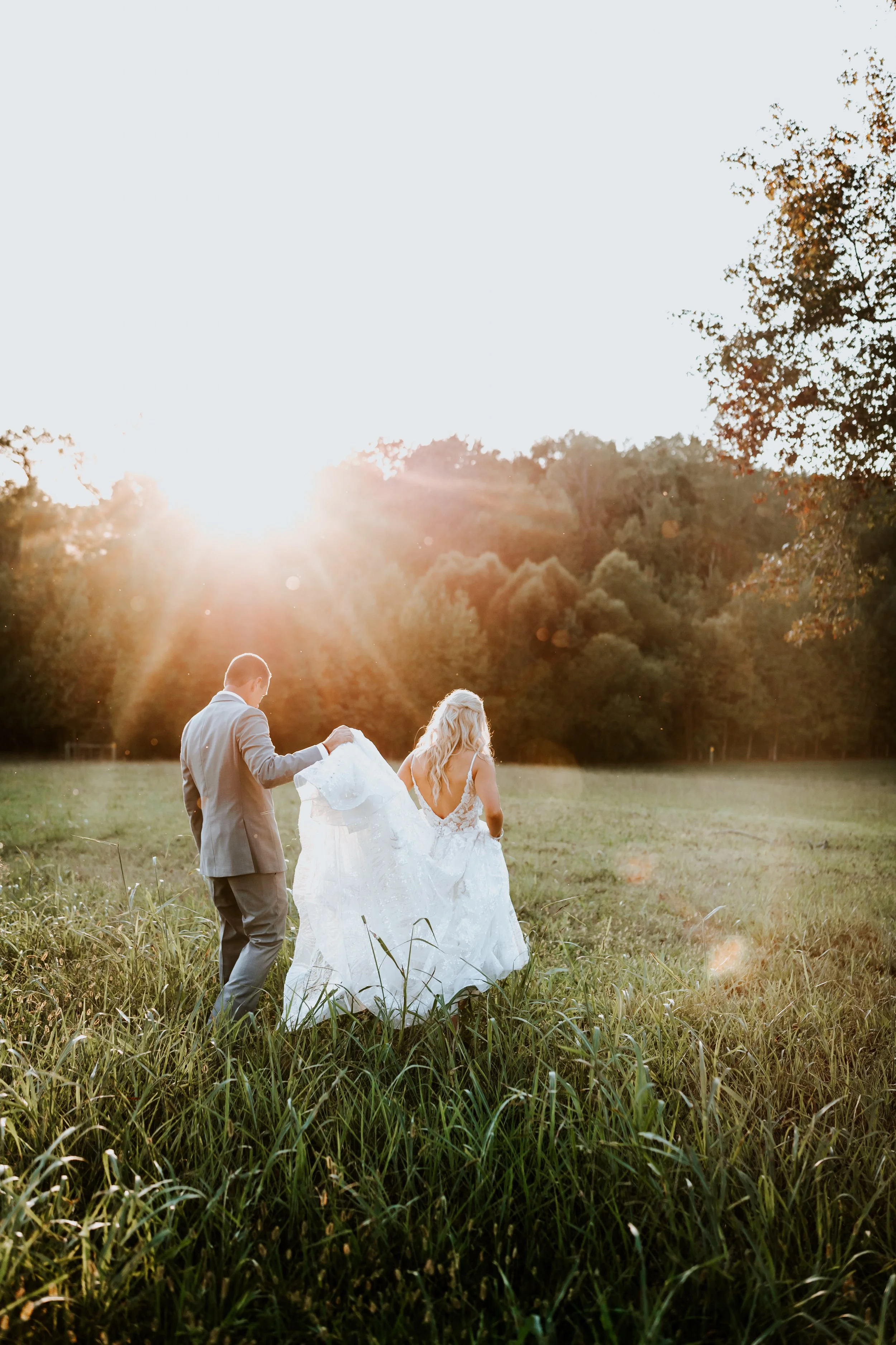 A wedding couple holding hands and walking through a grassy field at sunset, with trees in the background.