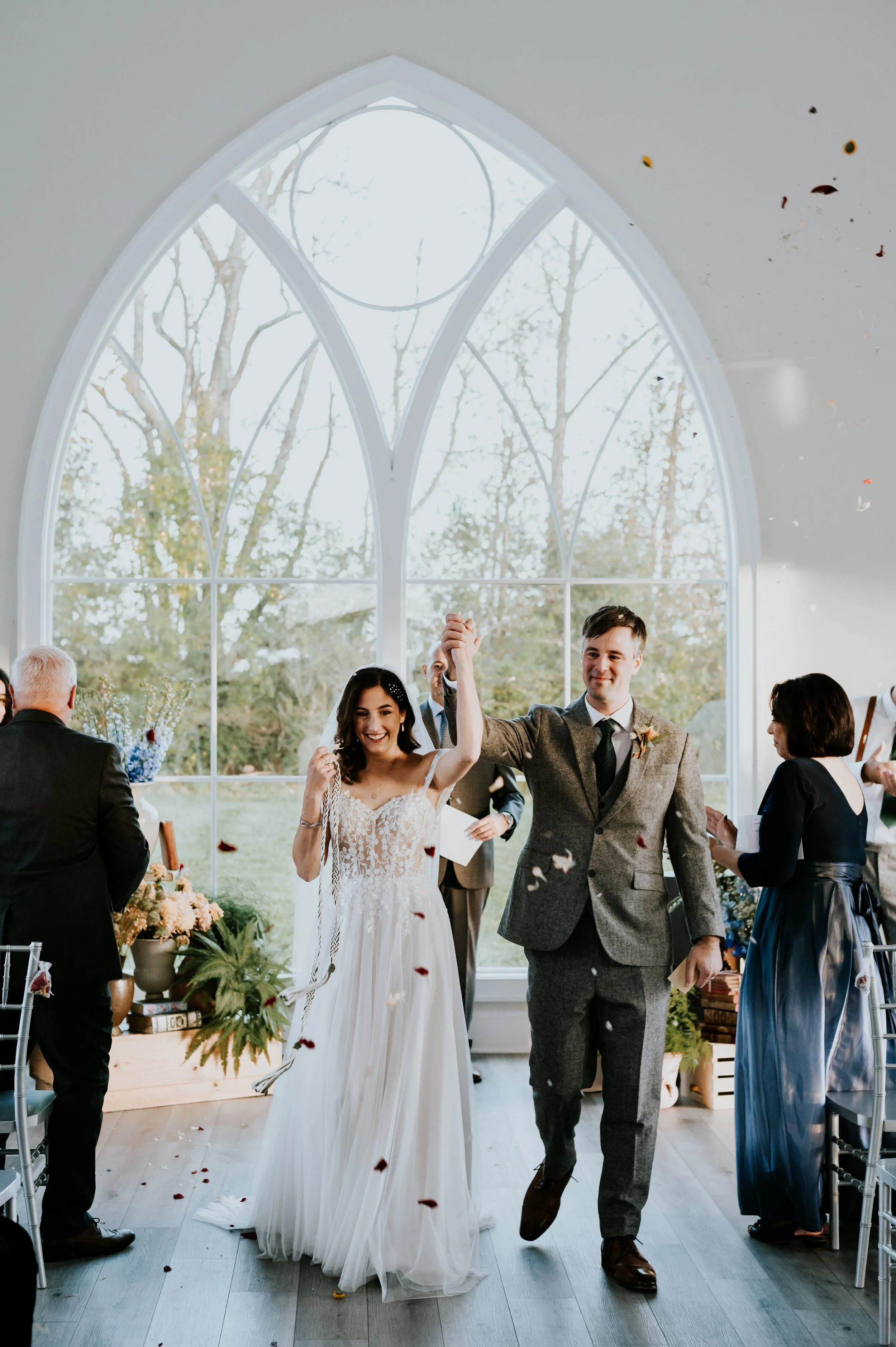 A bride and groom celebrating their wedding, holding hands and smiling, while walking through a reception hall filled with family and friends, with autumn trees visible through large arched windows.
