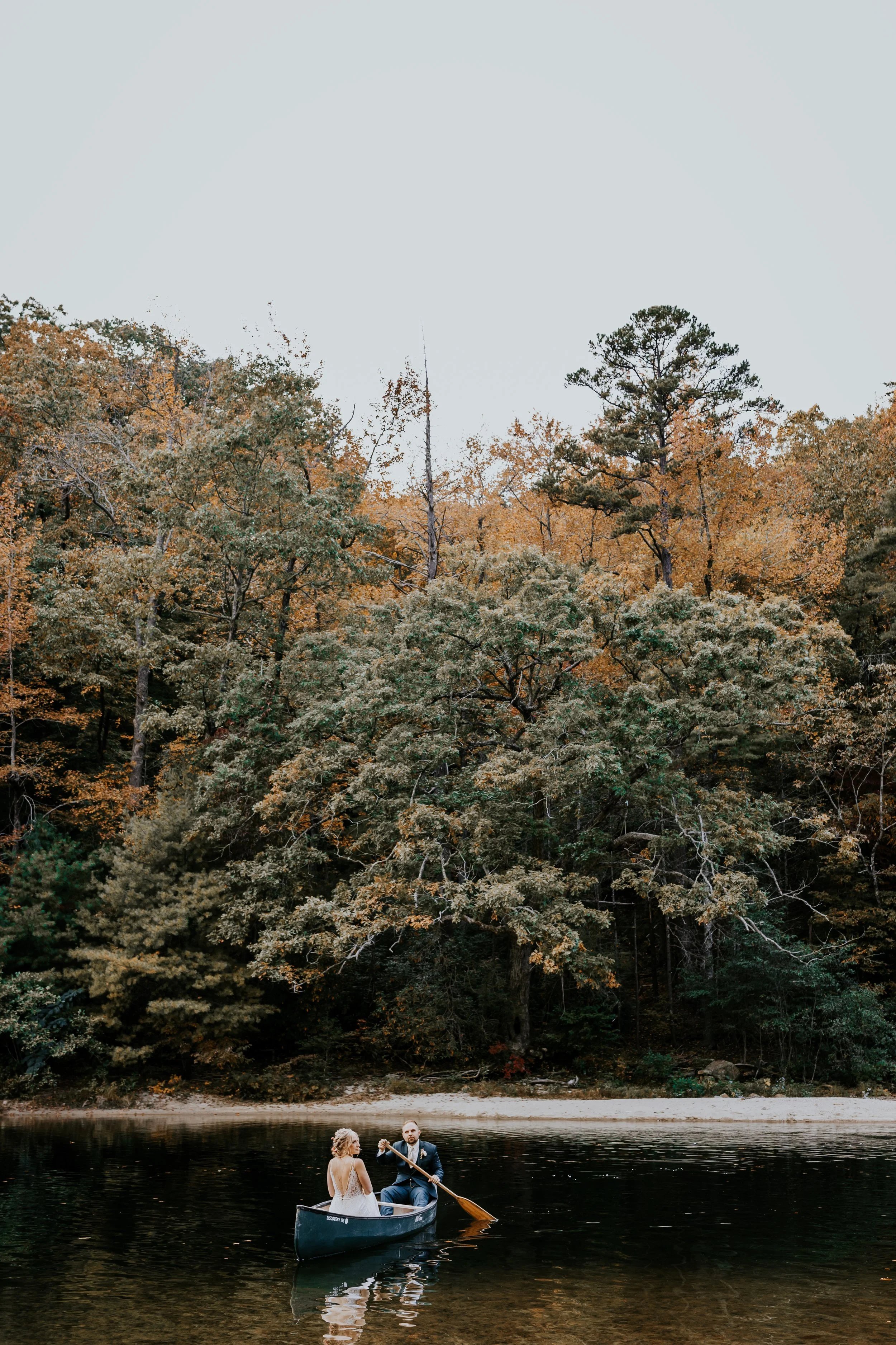 A couple in wedding attire in a small boat on a calm lake, surrounded by a forest with autumn-colored trees.