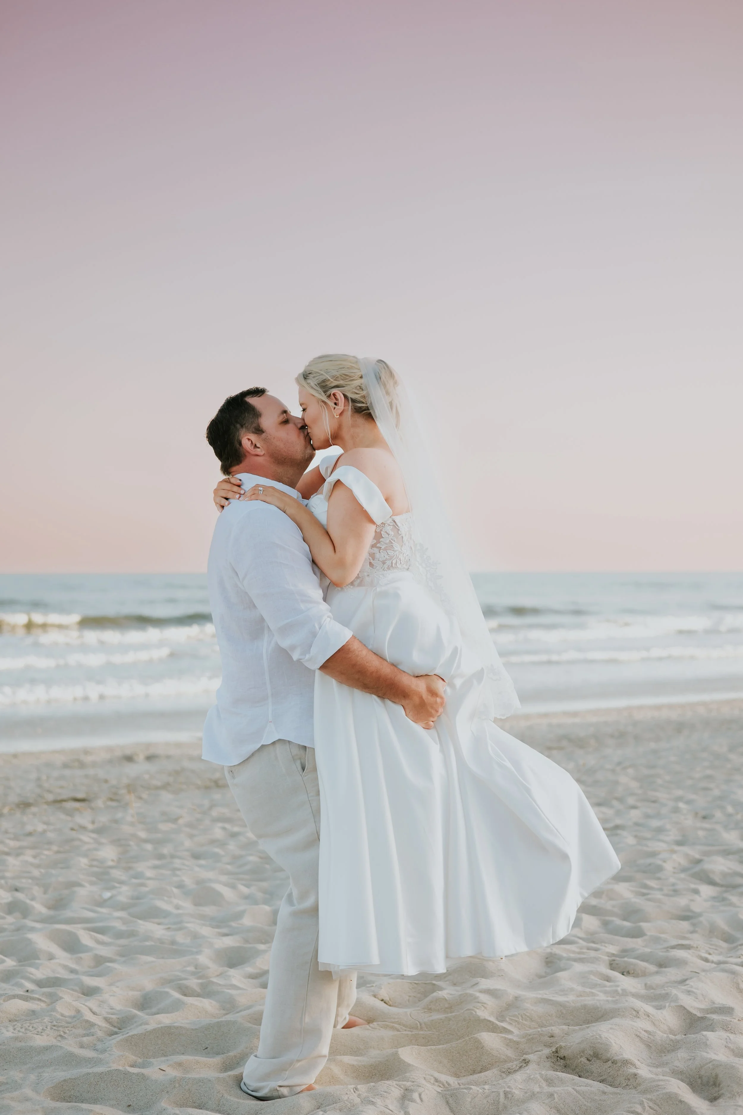 A couple, dressed in wedding attire, sharing a kiss on the beach during sunset, with the ocean and sky in the background.