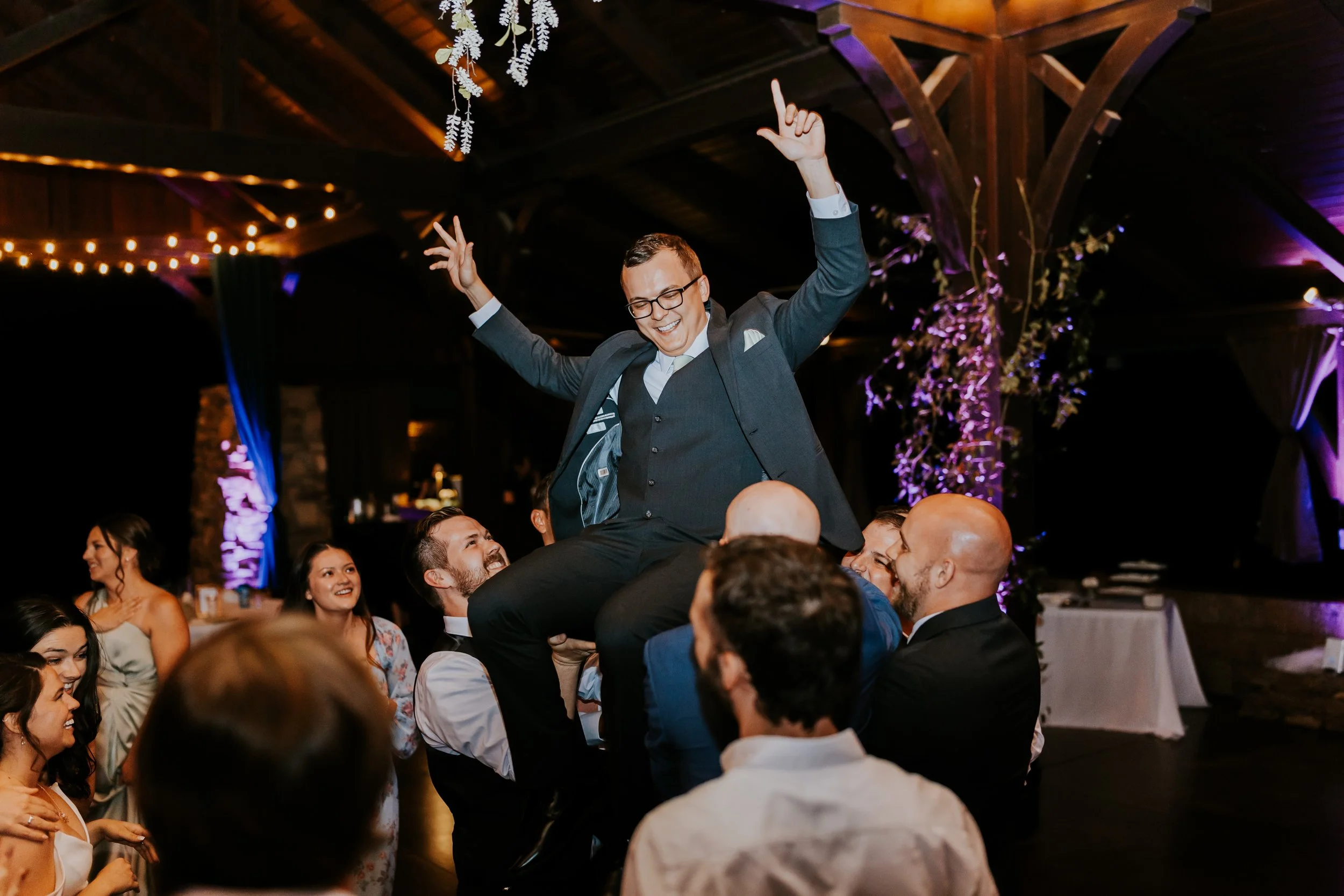 A man in a gray suit and glasses, smiling and holding his arms up, is lifted on the shoulders of a group of people at a lively celebration in a decorated indoor venue with warm lighting and hanging floral decorations.